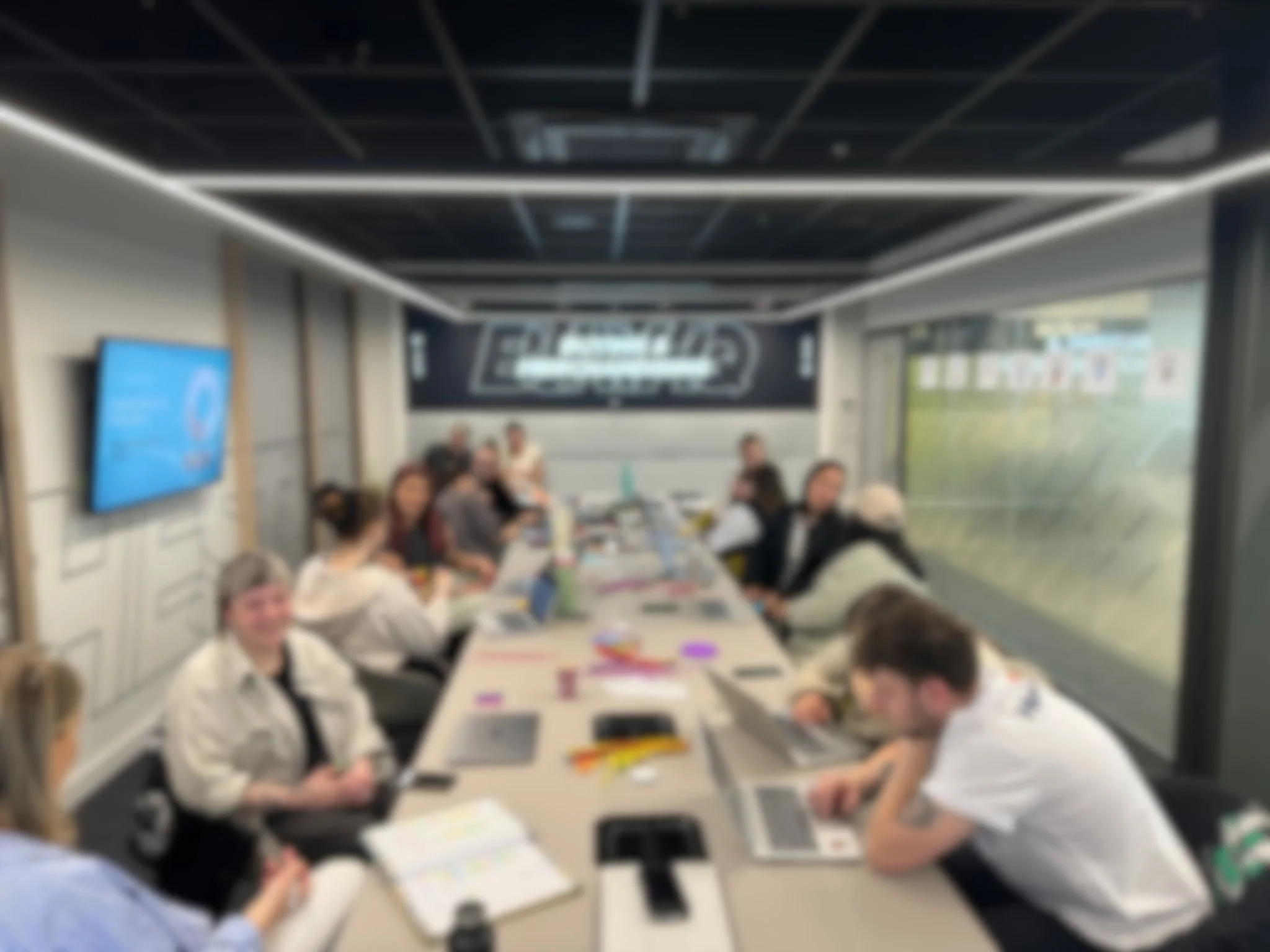 A group of people sitting around a long conference table in a modern office space, some working on laptops, with a large screen on the wall displaying a presentation.