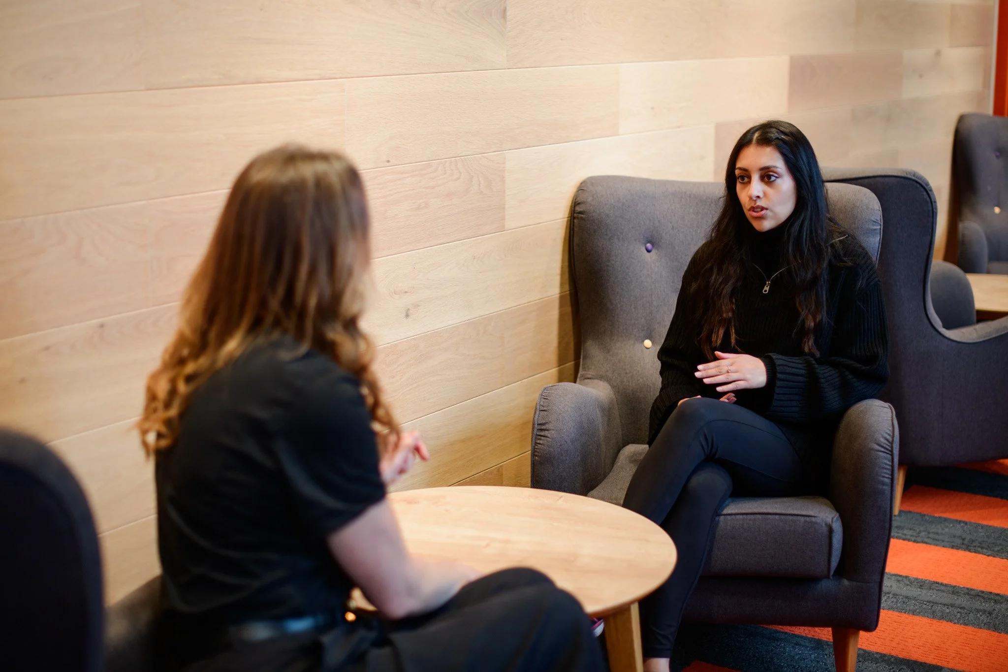Two women are having a conversation in a cozy, modern lounge area. One woman with long dark hair is sitting in a cushioned armchair, facing the other woman with lighter hair seated at a small round table.