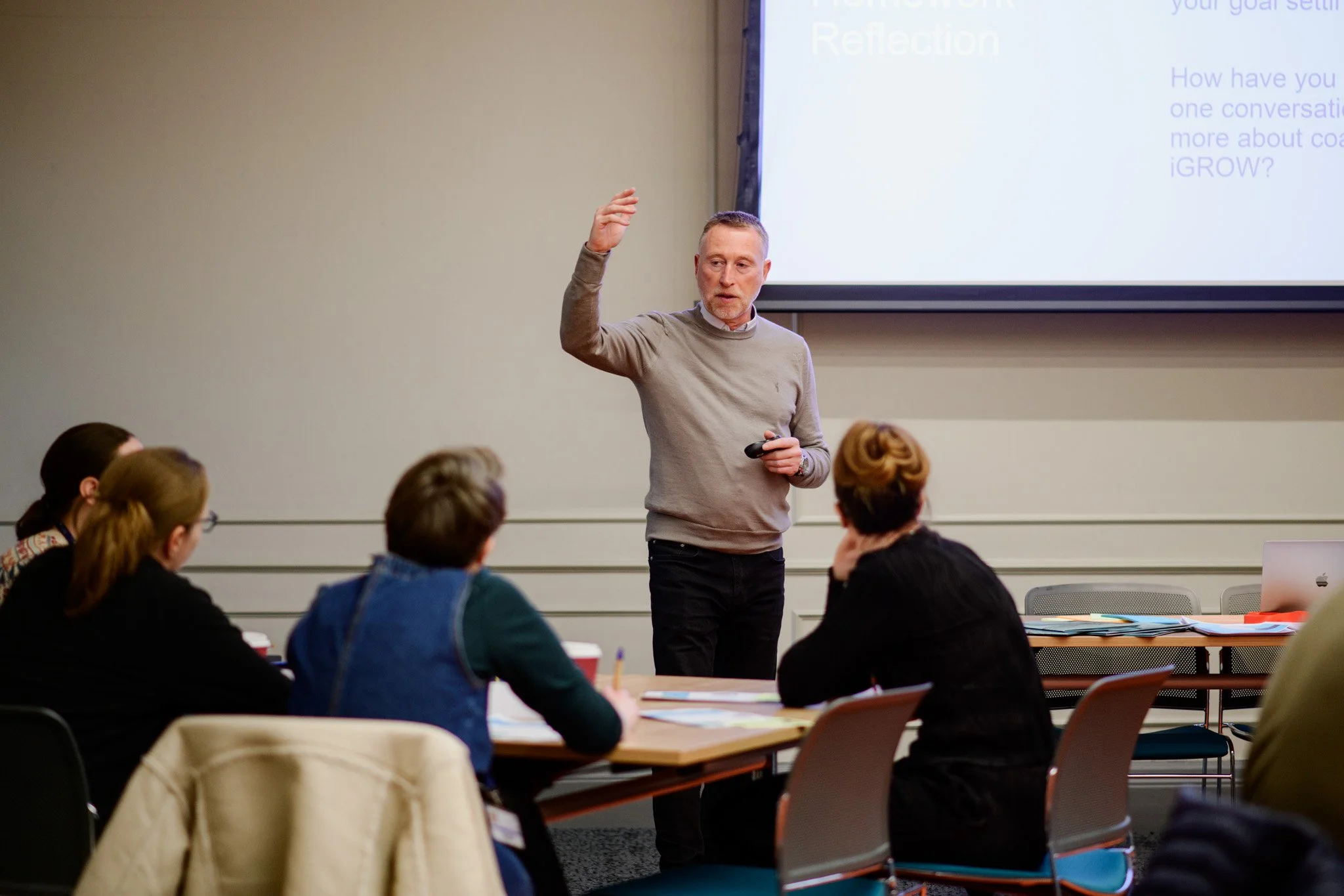 A man standing and giving a presentation in a classroom or conference room with a large projection screen behind him, addressing a group of seated women students or professionals.