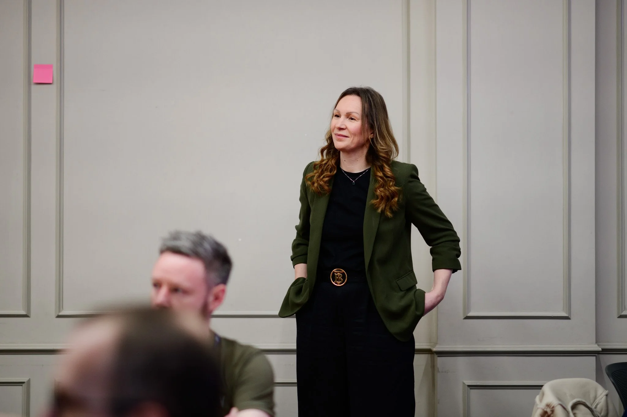 A woman with long, wavy brown hair wearing a dark green blazer and black shirt, standing in a meeting room with beige walls and decorative panels, with her hands in her pockets, smiling slightly.