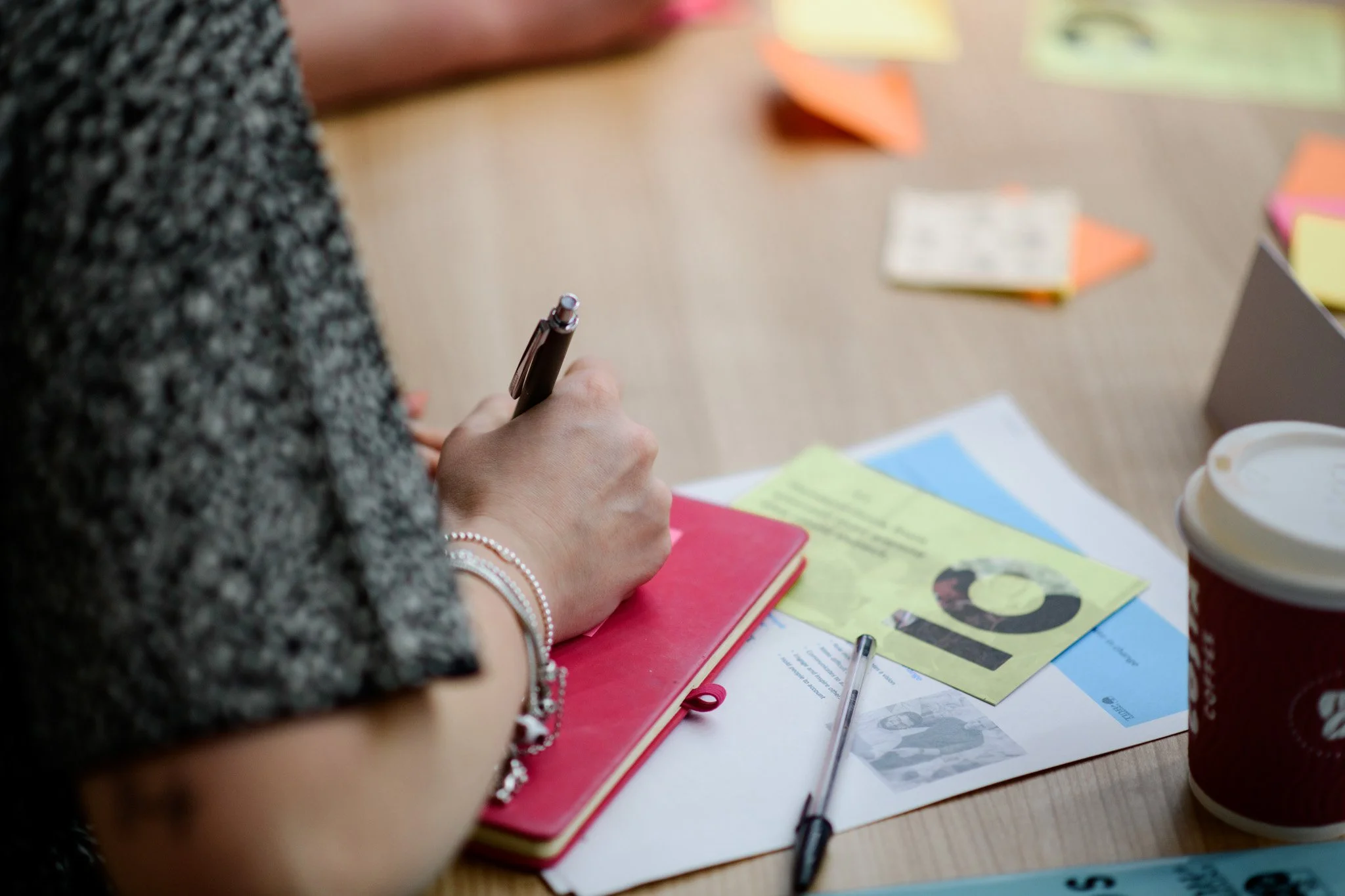 A person writing in a red notebook surrounded by papers, a pen, colorful sticky notes, and a coffee cup on a wooden table.