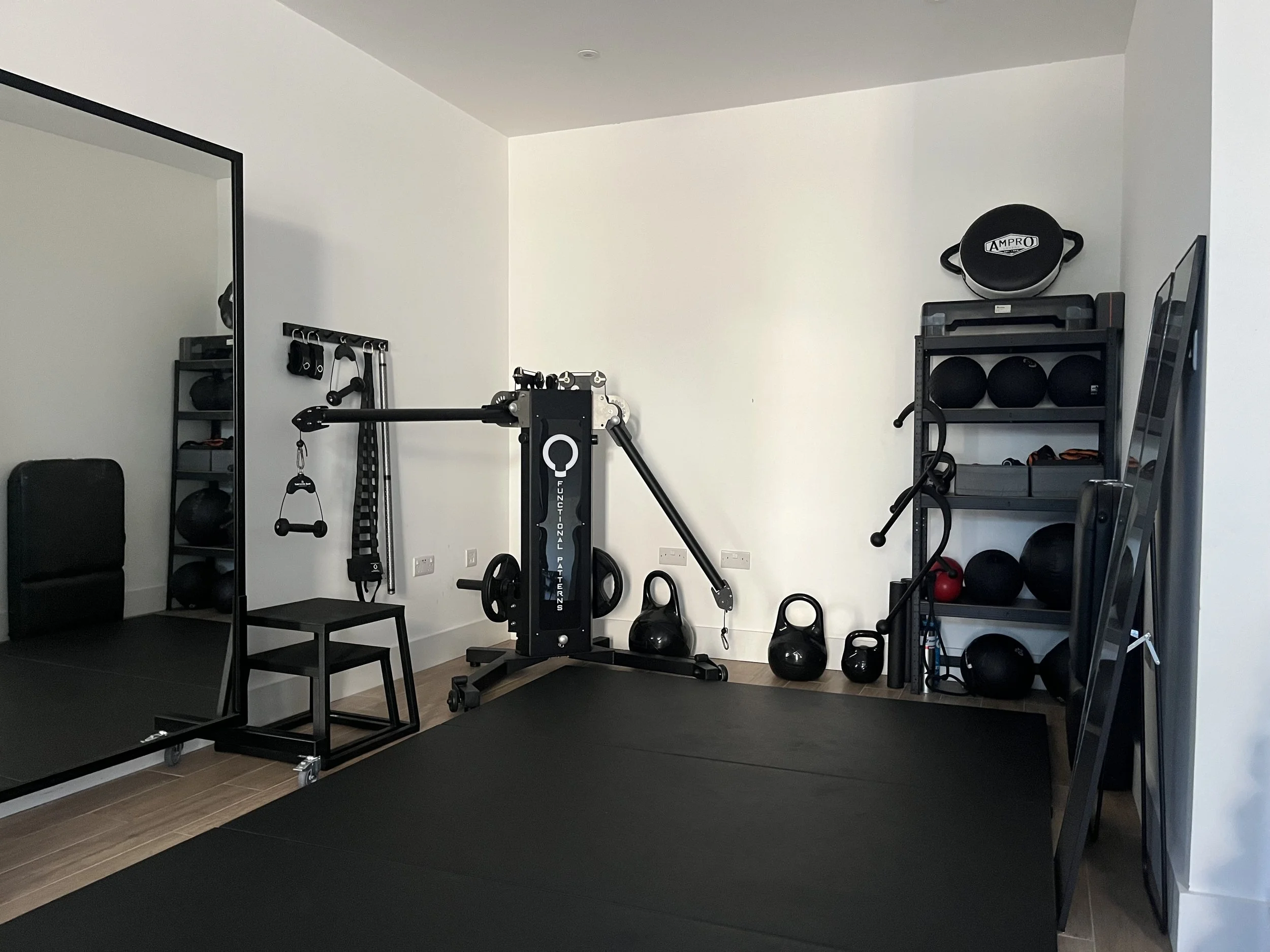 London functional gym with black flooring, a large mirror on the left, displaying exercise equipment including kettlebells, medicine balls, and a wall-mounted rack holding various workout gear, and a black Functional Patterns pulley machine.