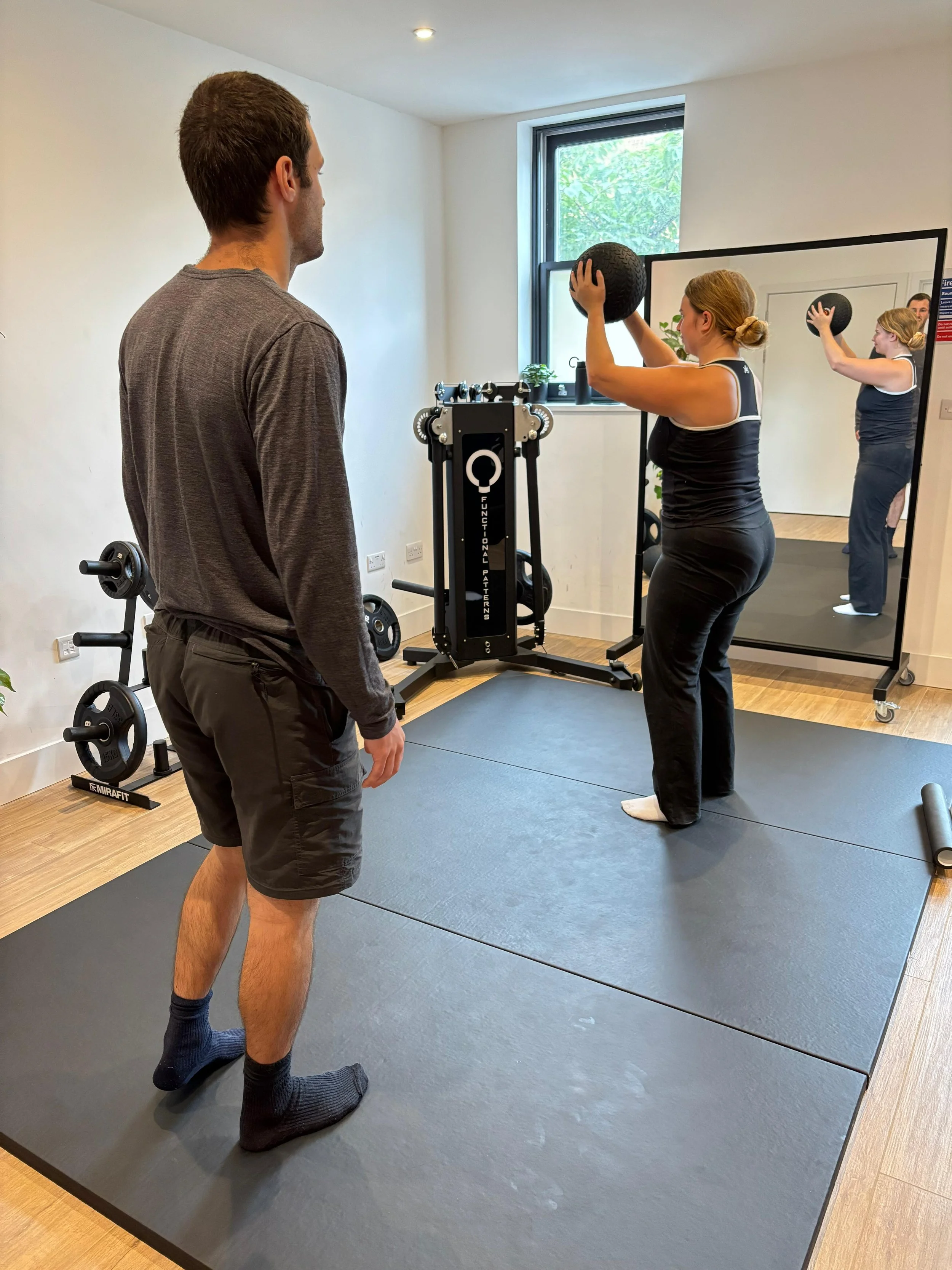 A woman exercising with a medicine ball in front of a mirror, while a Functional Patterns trainer observes her in a London fitness studio. The woman is preparing to throw the ball, and her reflection shows her from behind.