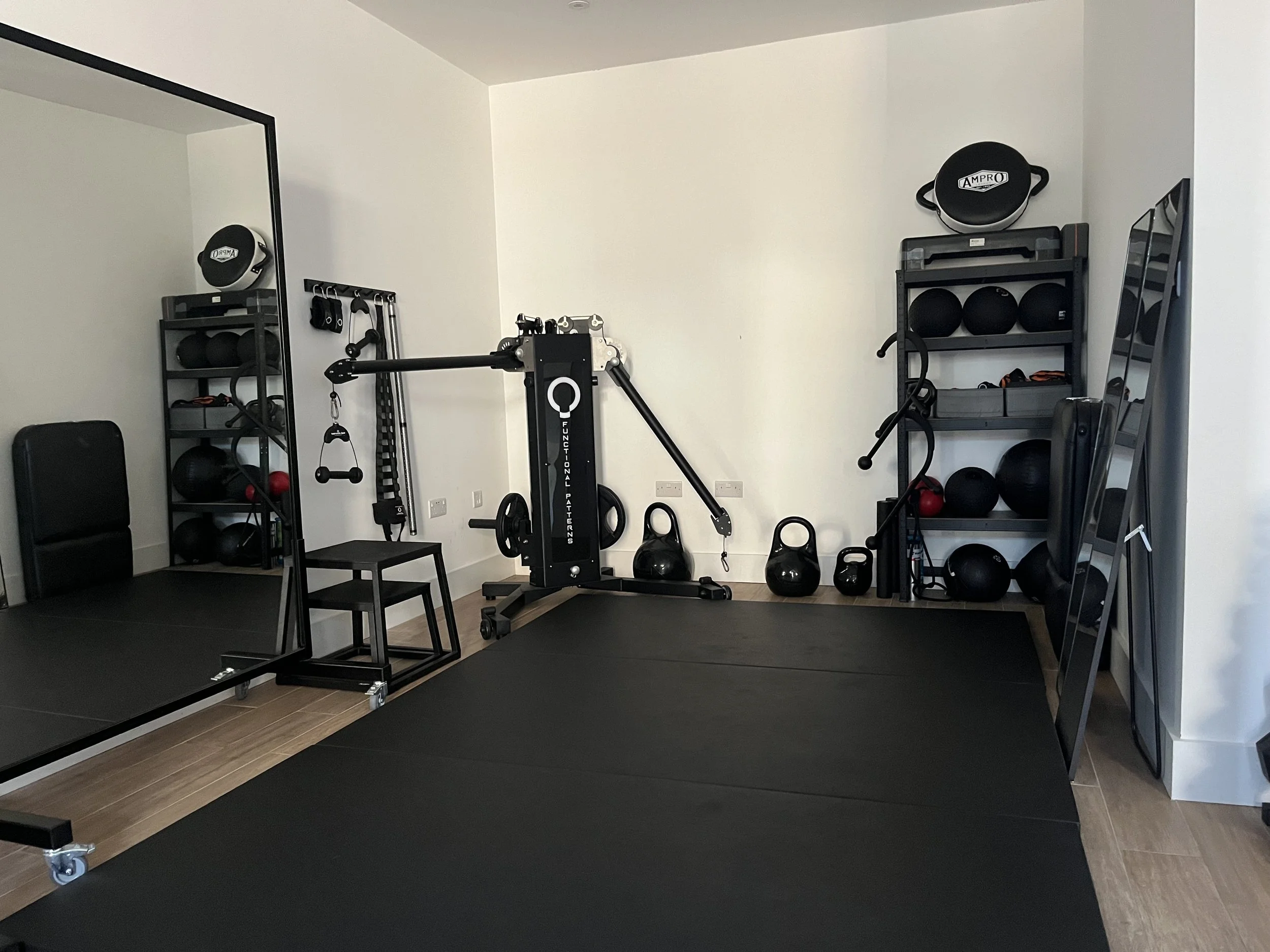 A Functional Patterns gym in London with black mats on the floor, shelving units with black and red exercise equipment, a large mirror, kettlebells, and a workout machine in a room with white walls.