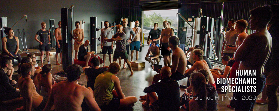 A Functional Patterns Human Biomechanics Specialist course with people sitting on the floor and standing, watching a trainer demonstrate Functional Patterns corrective exercises in a gym. The gym has large windows and workout equipment.
