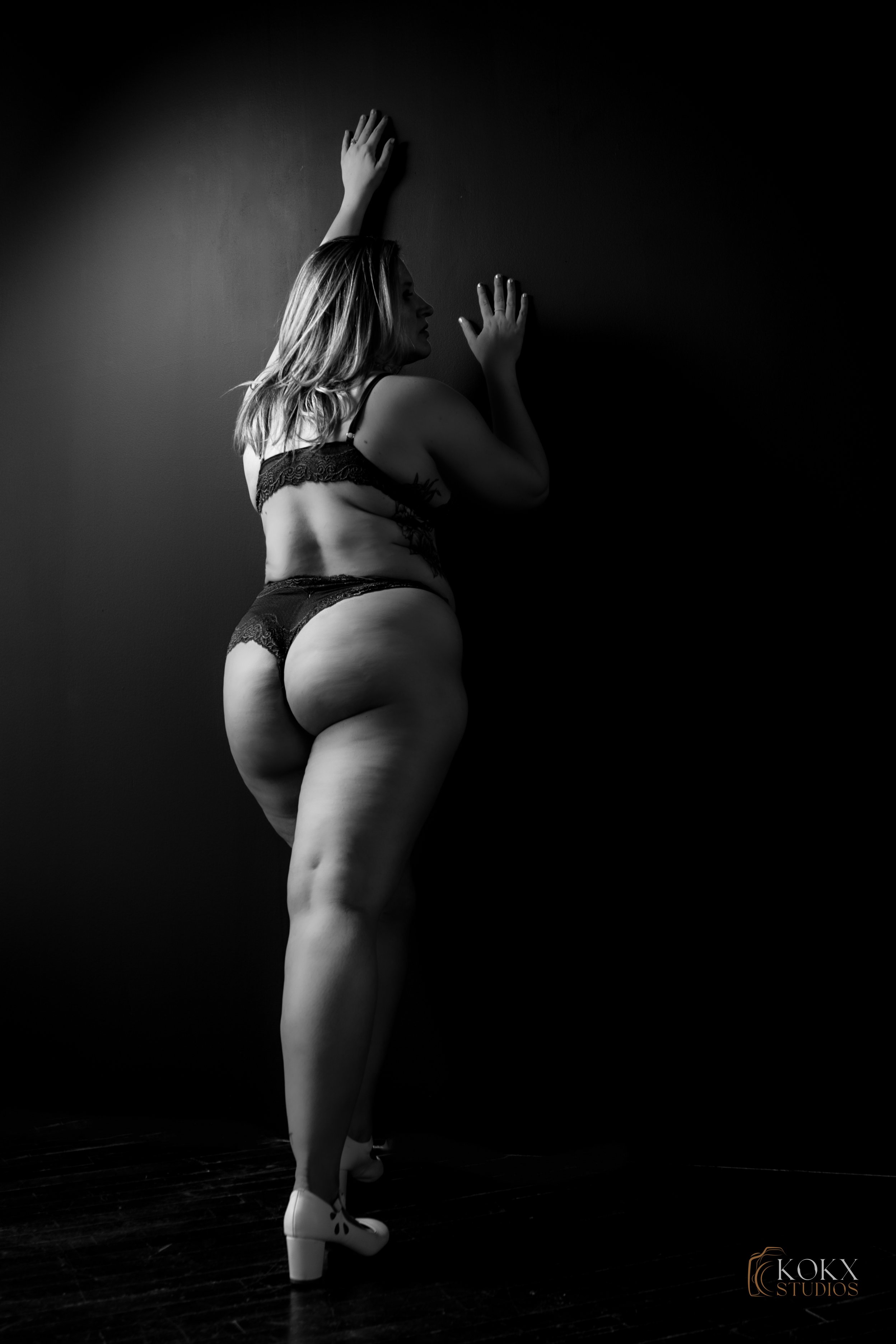 A woman in lingerie and white high heels poses with her back to the camera against a dark wall.