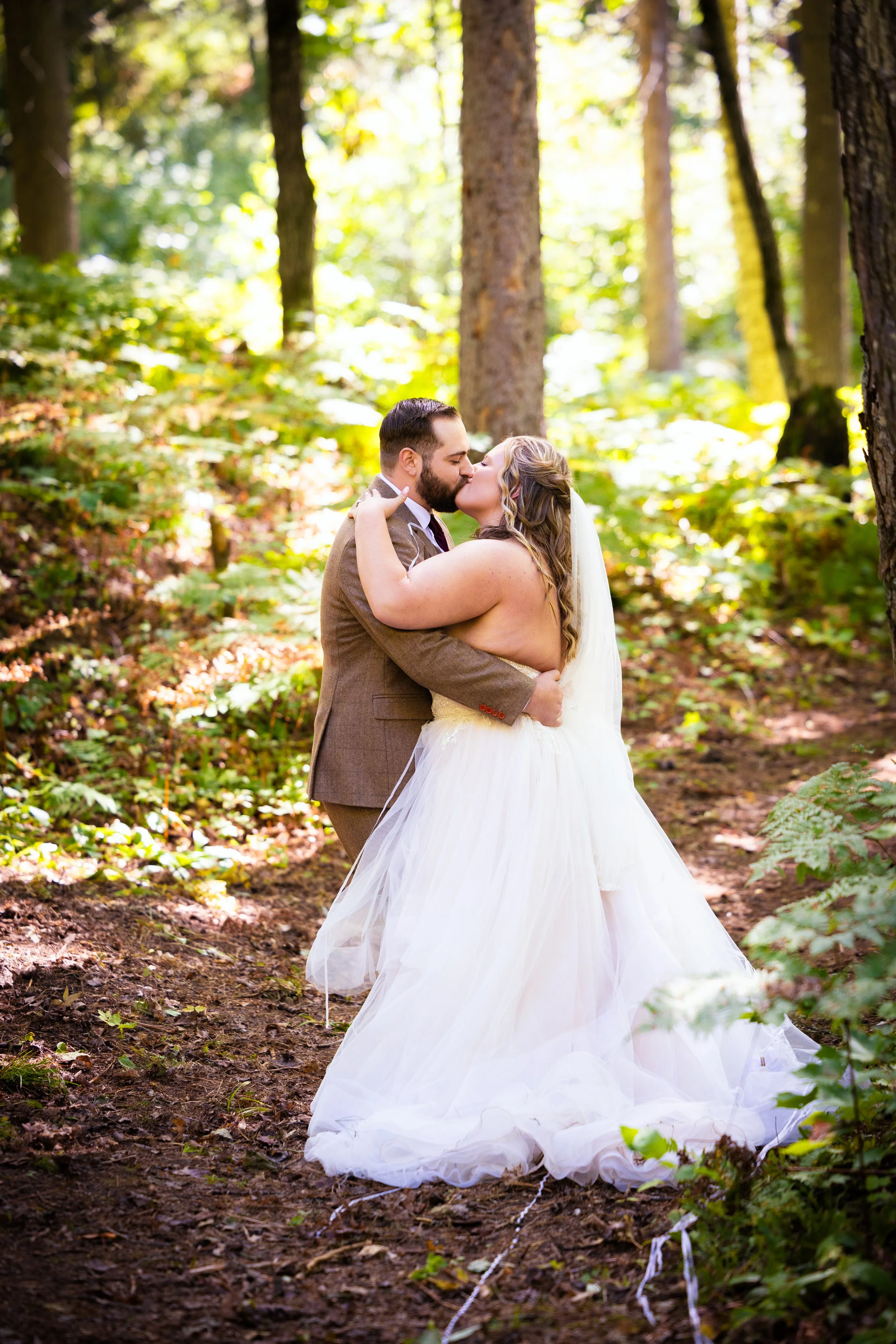 A bride and groom kiss in a forest clearing surrounded by green trees and sunlight filtering through.