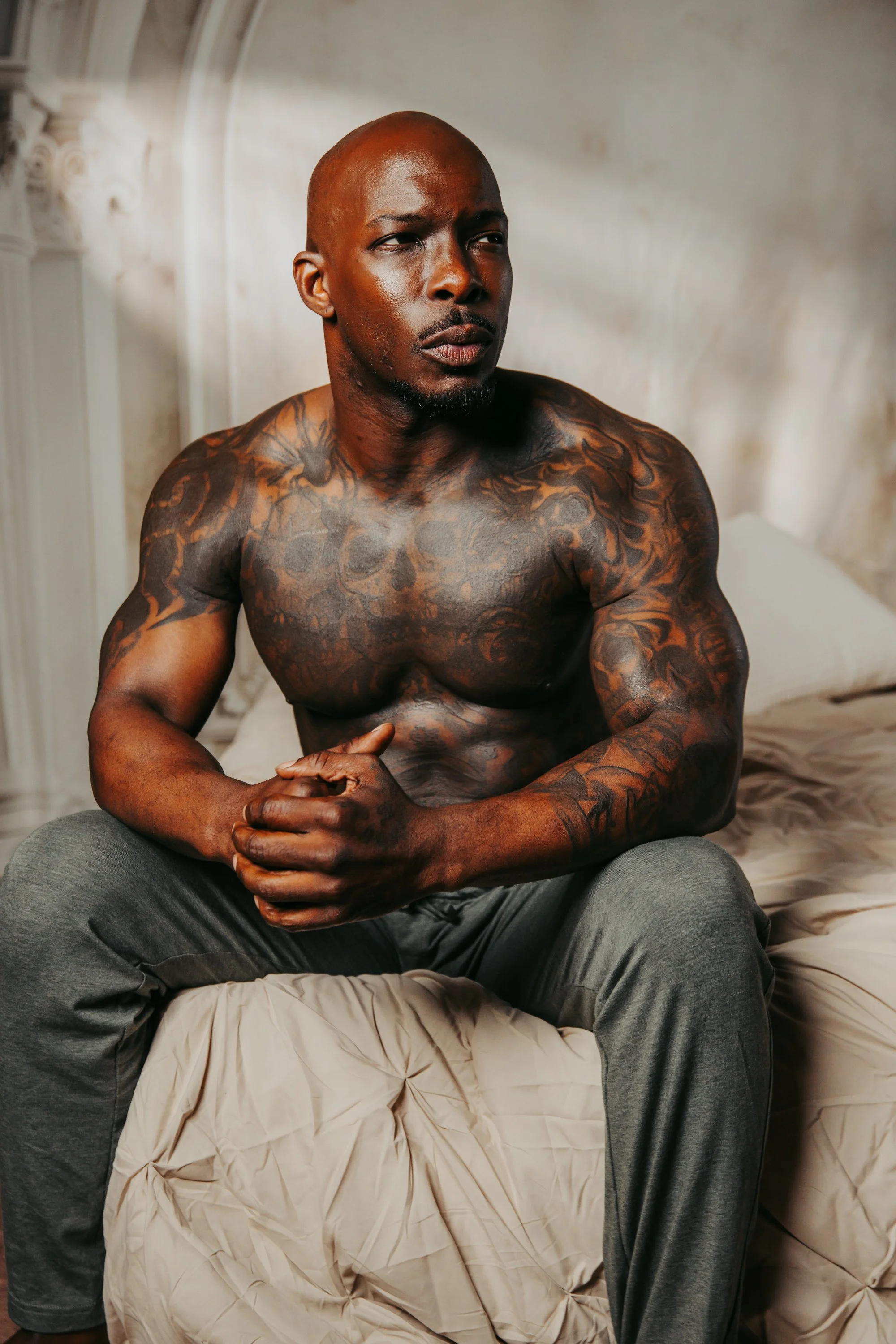 A shirtless man with tattoos sitting on a bed in a room with cream-colored walls.