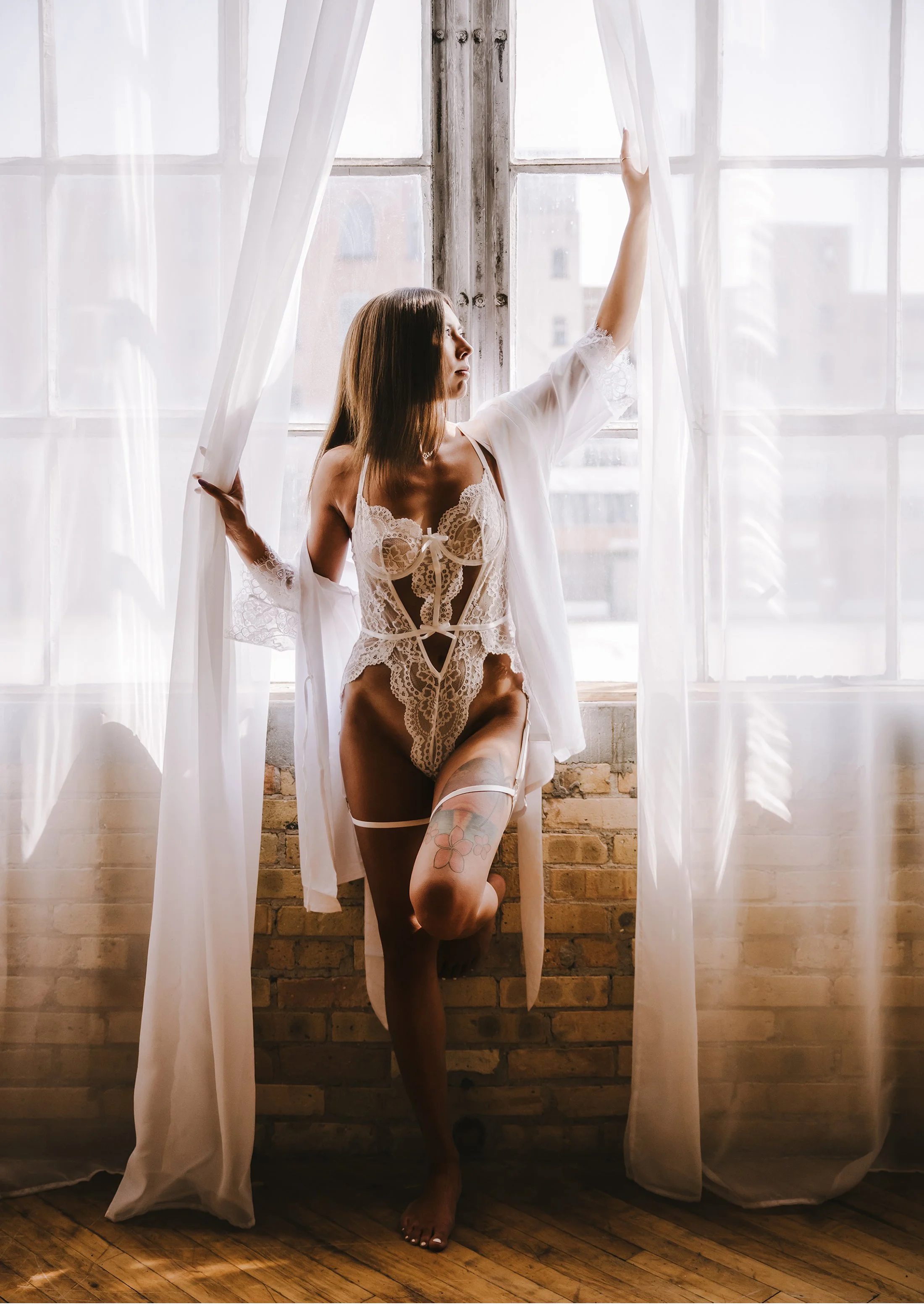 Woman in lace lingerie and white robe standing by a large window with sheer curtains, looking outside.