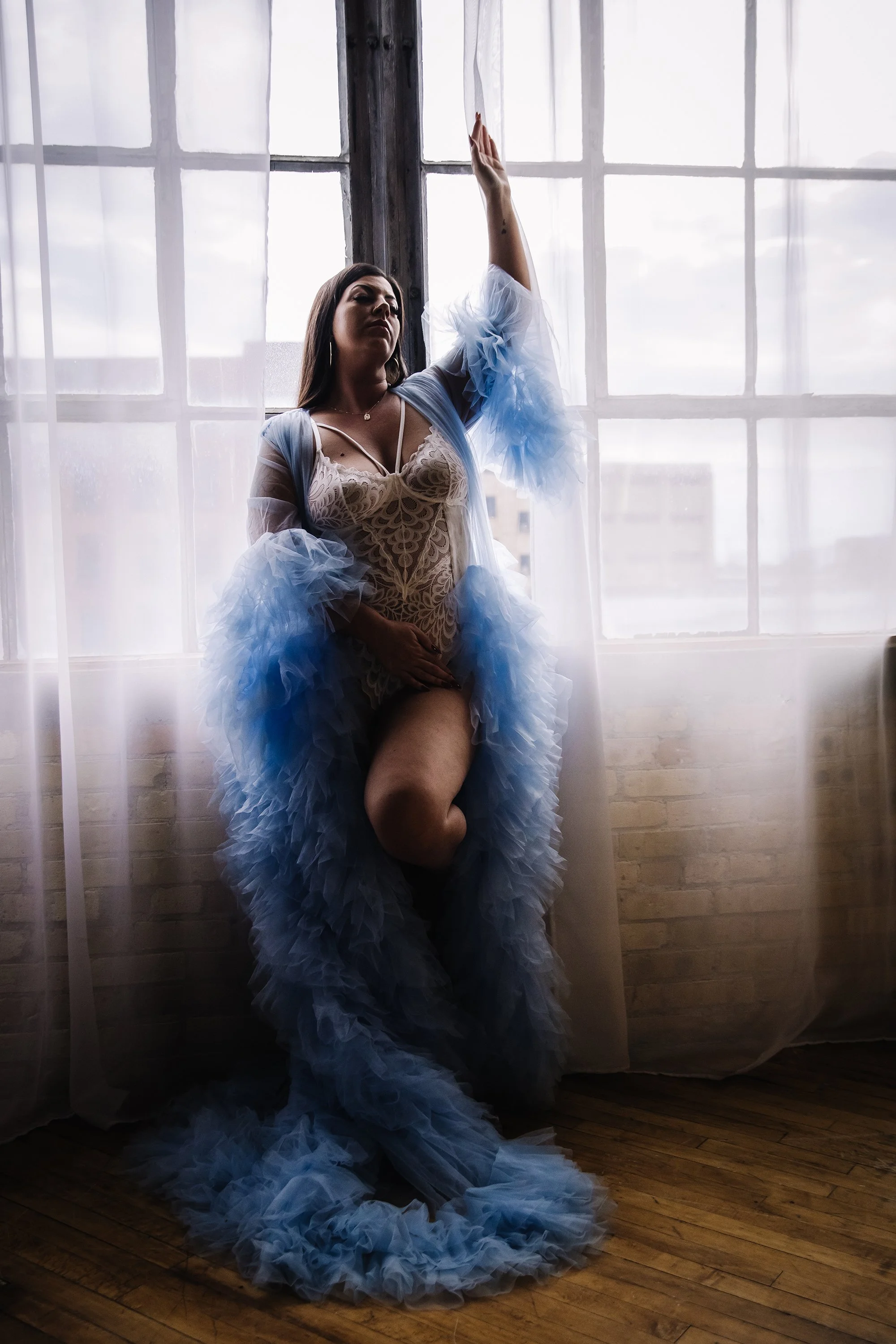 A woman standing by a window in a sheer dress with puffy blue tulle around her arms and legs, posing with one arm raised.
