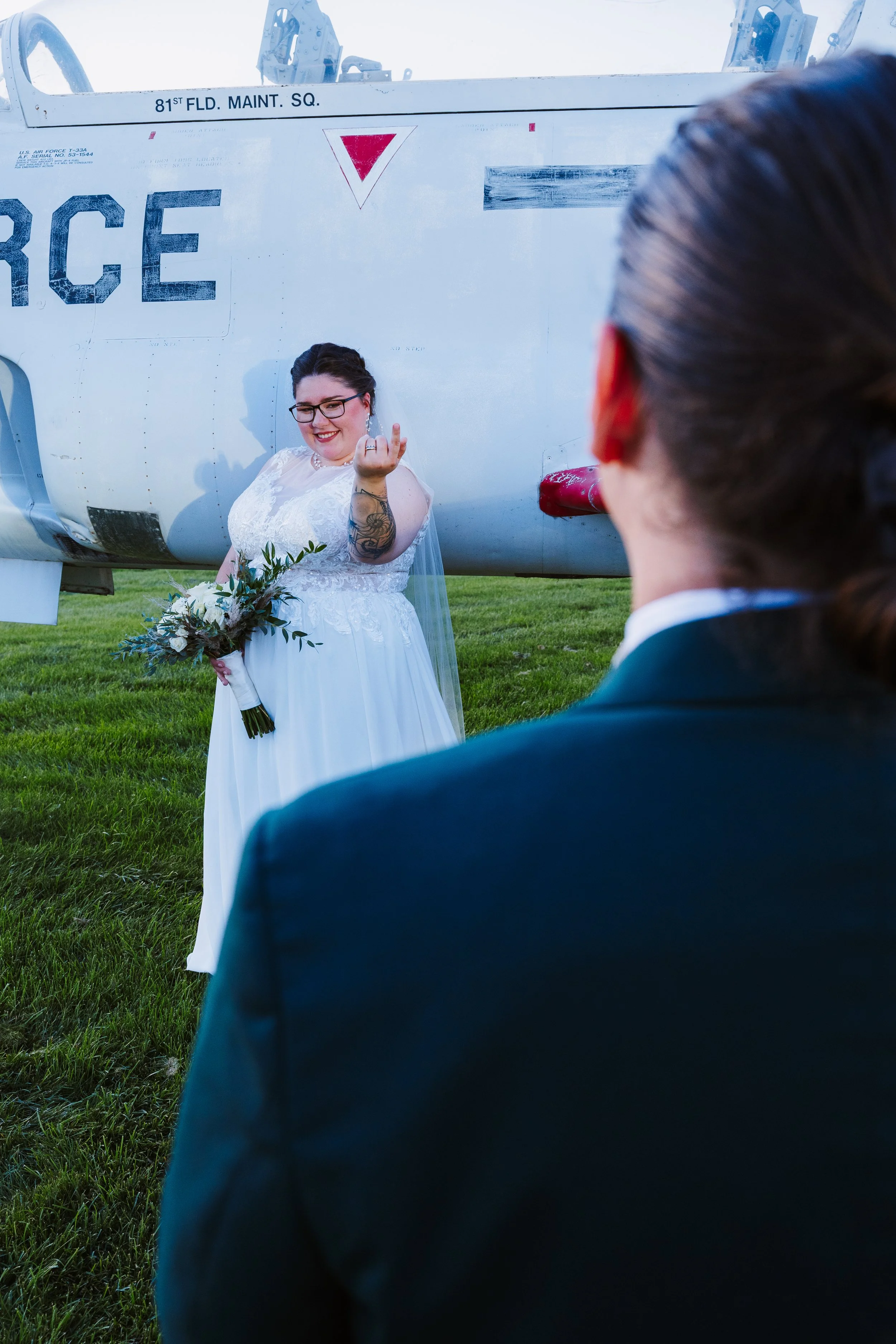 A bride in a white dress and glasses holding a bouquet, making a rude hand gesture towards a person in a suit standing in front of her outdoors on a grassy area, with a military helicopter in the background.