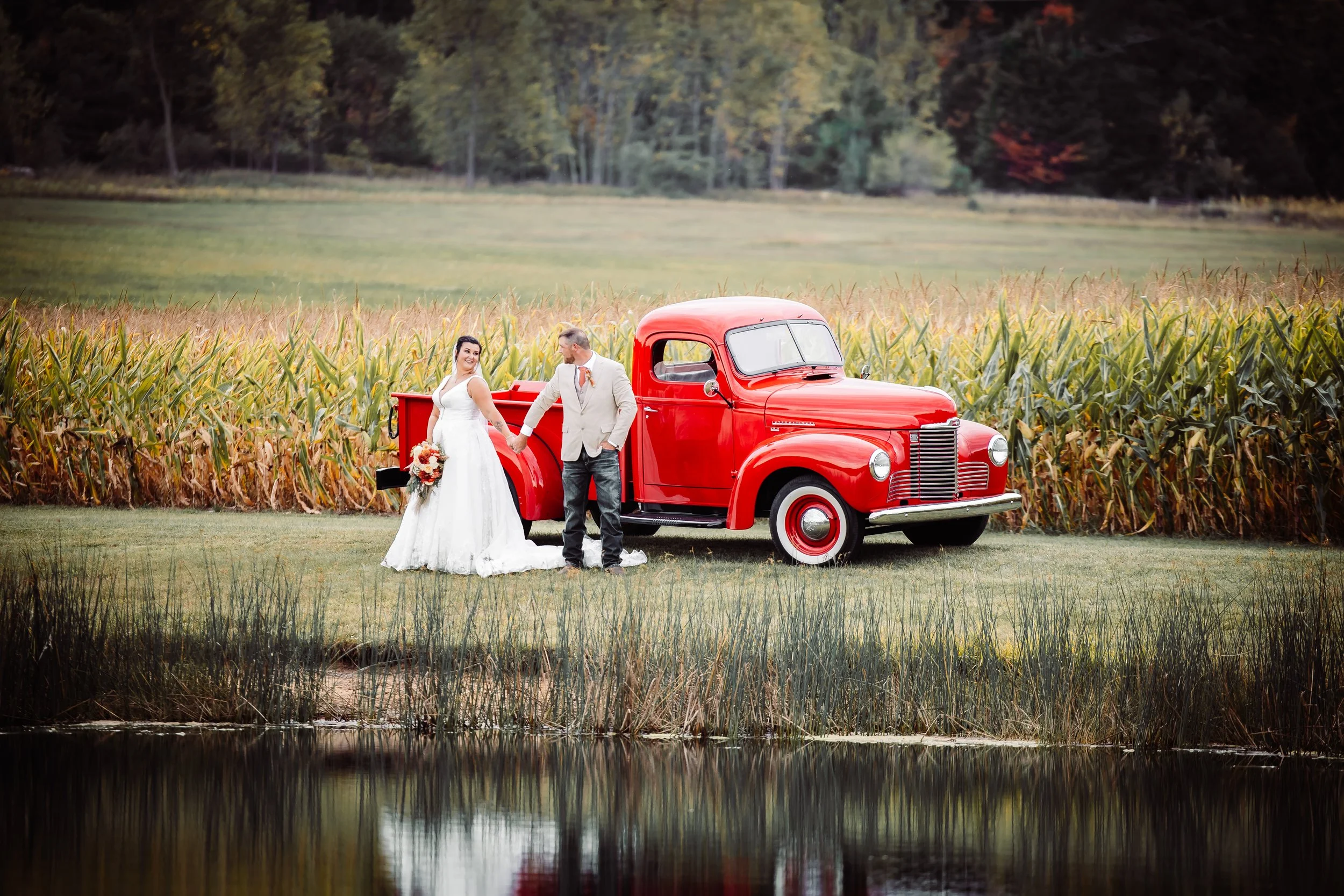 A bride and groom standing beside a red vintage truck in a field of tall corn, with a pond in the foreground and a wooded area in the background.