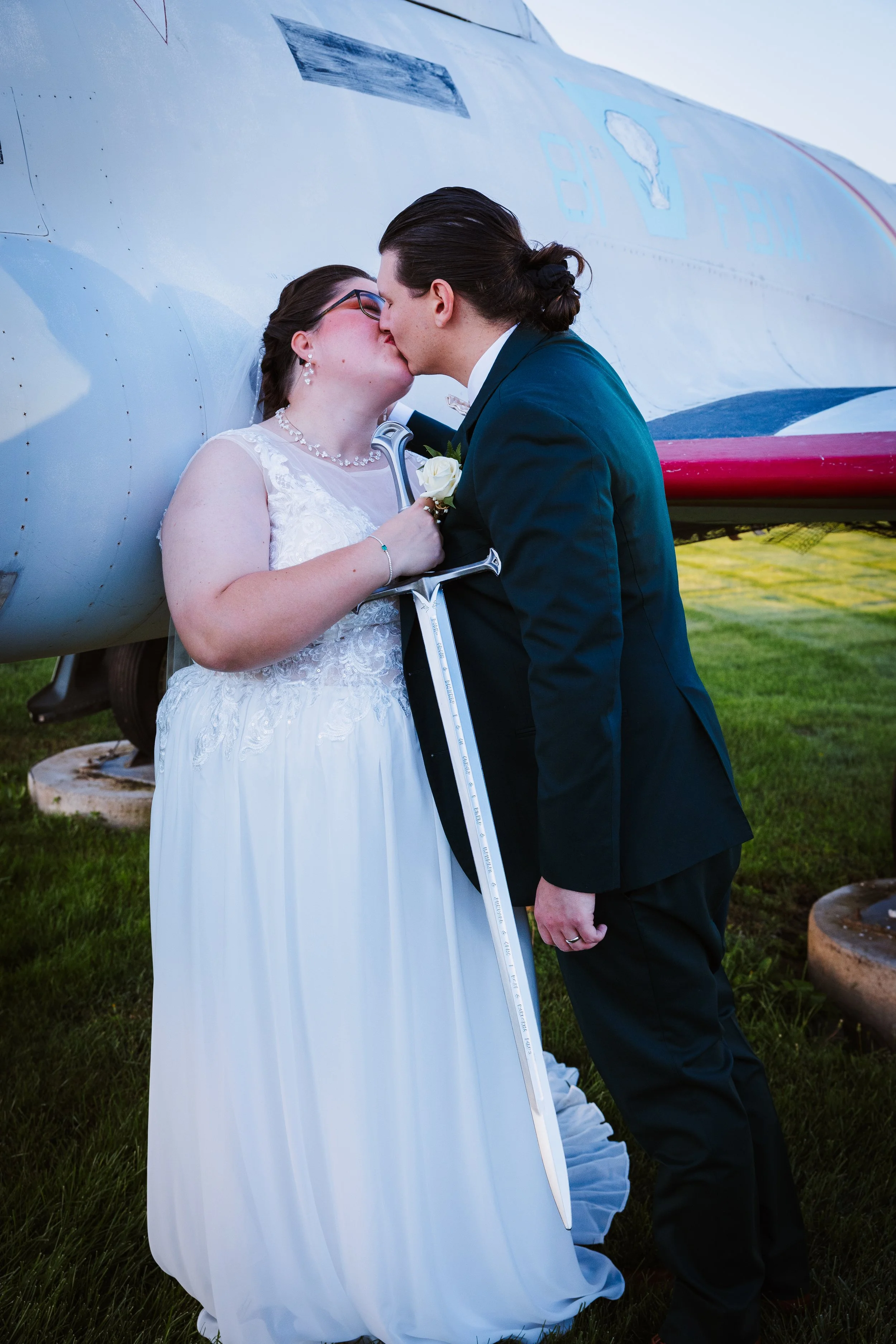A couple in wedding attire sharing a kiss, standing outdoors in front of an airplane. The bride is holding a sword and a white rose, and the groom is dressed in a dark suit with his hair tied back.
