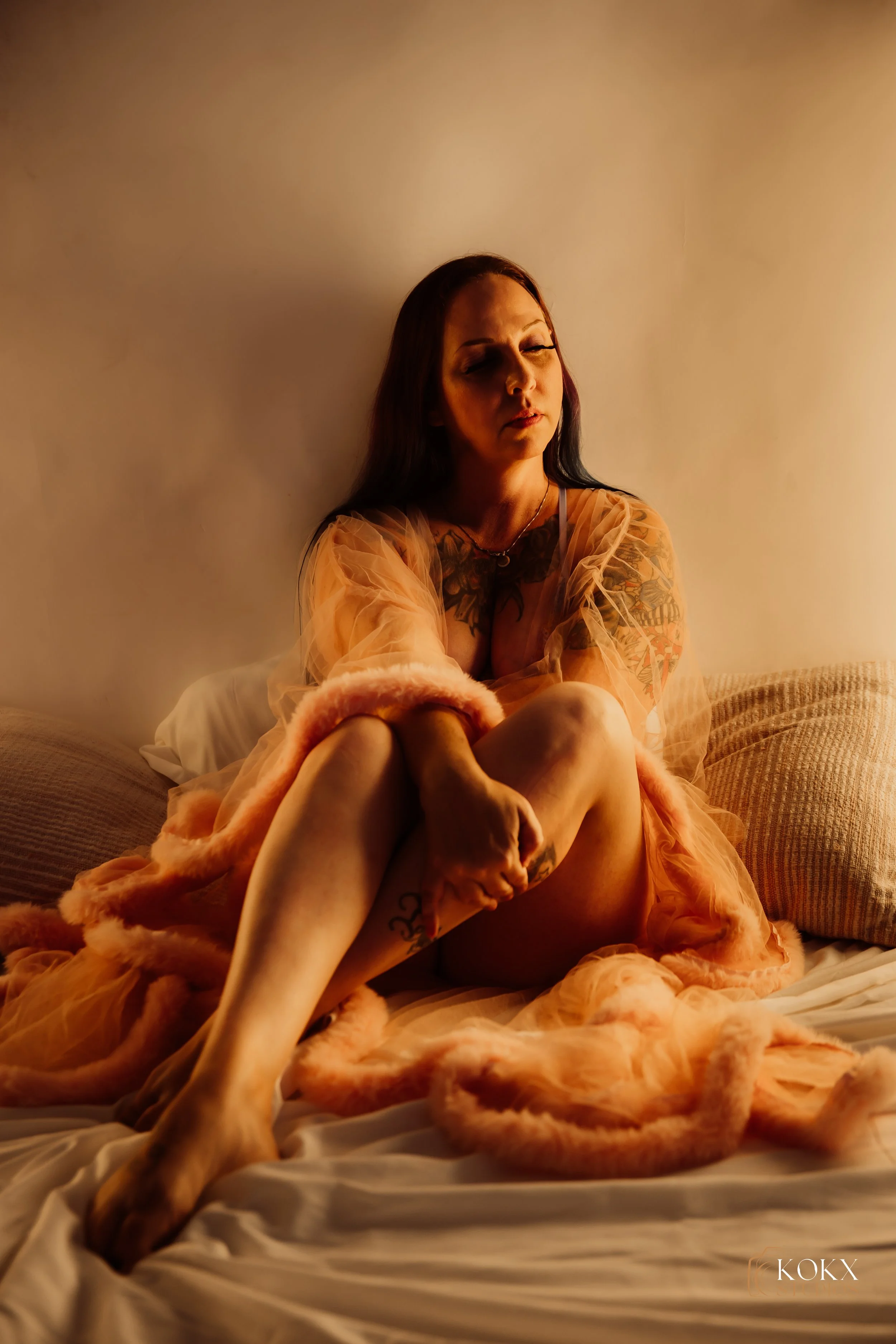 A woman with tattoos sitting on a bed, wrapped in a peach-colored fuzzy robe, with a neutral expression and her eyes partially closed.