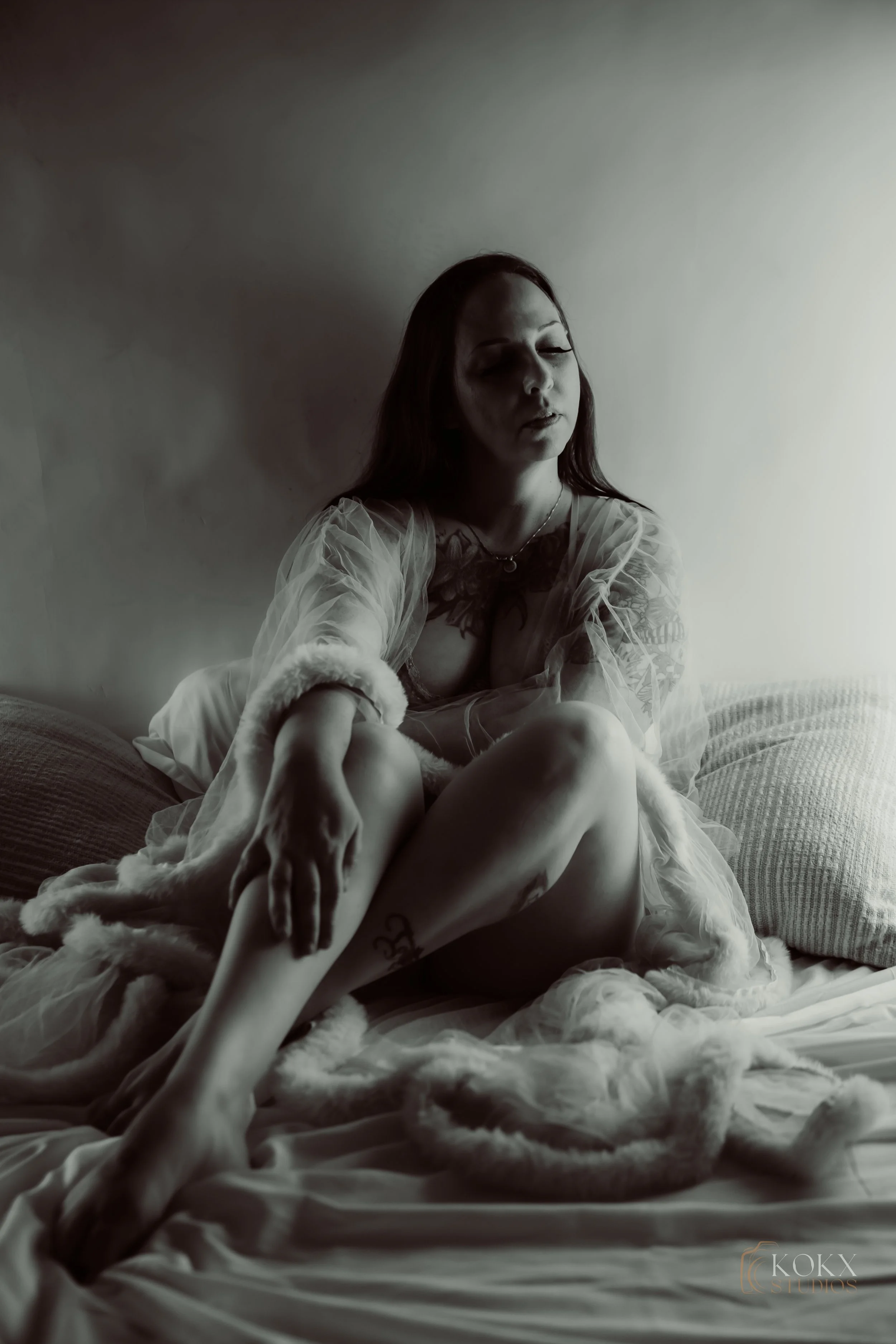 A black-and-white photo of a woman sitting on a bed, wearing a sheer robe with fur trim, with her eyes closed and a contemplative expression.