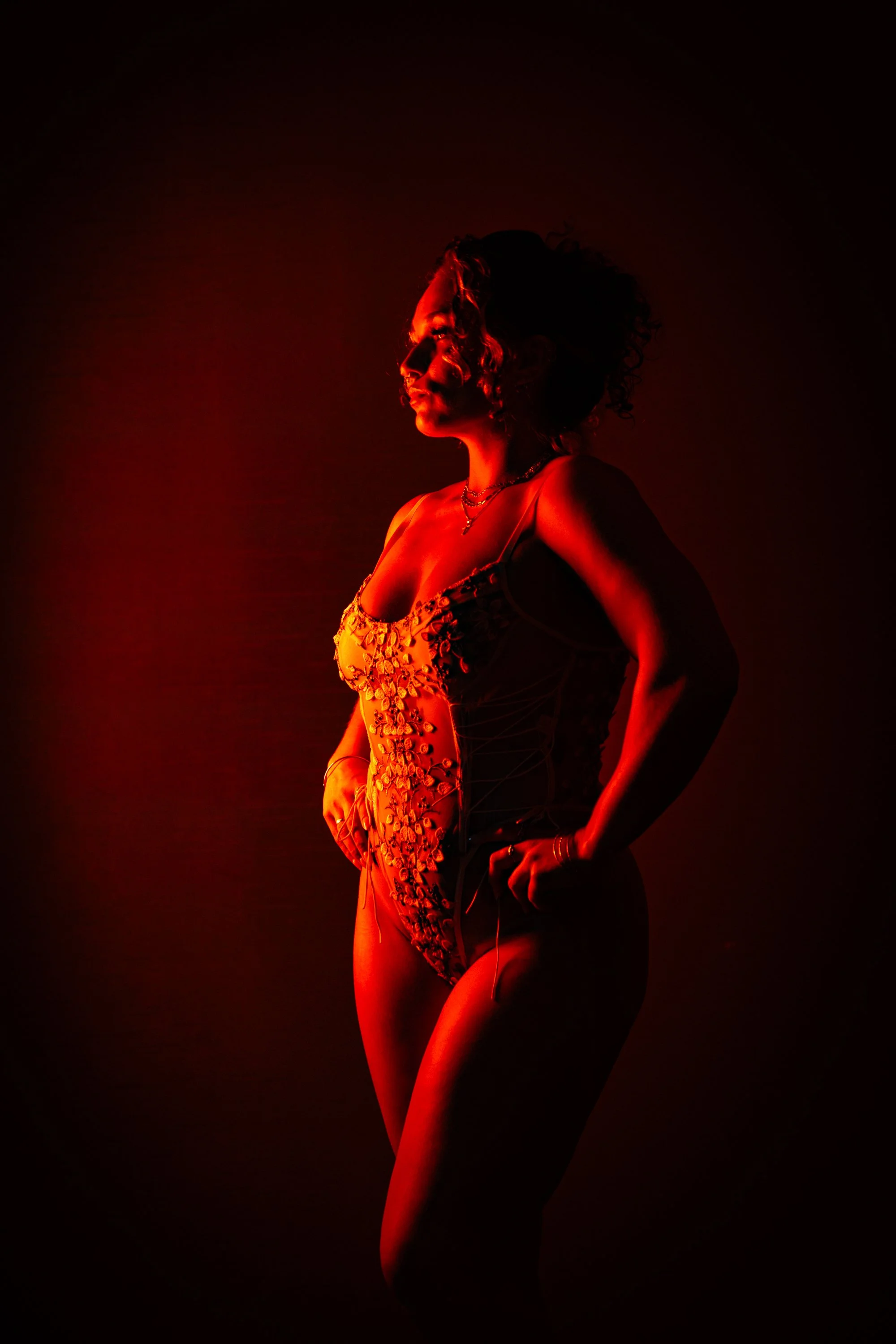 A woman posing in a dark setting illuminated by red light, wearing a revealing floral embroidered outfit.