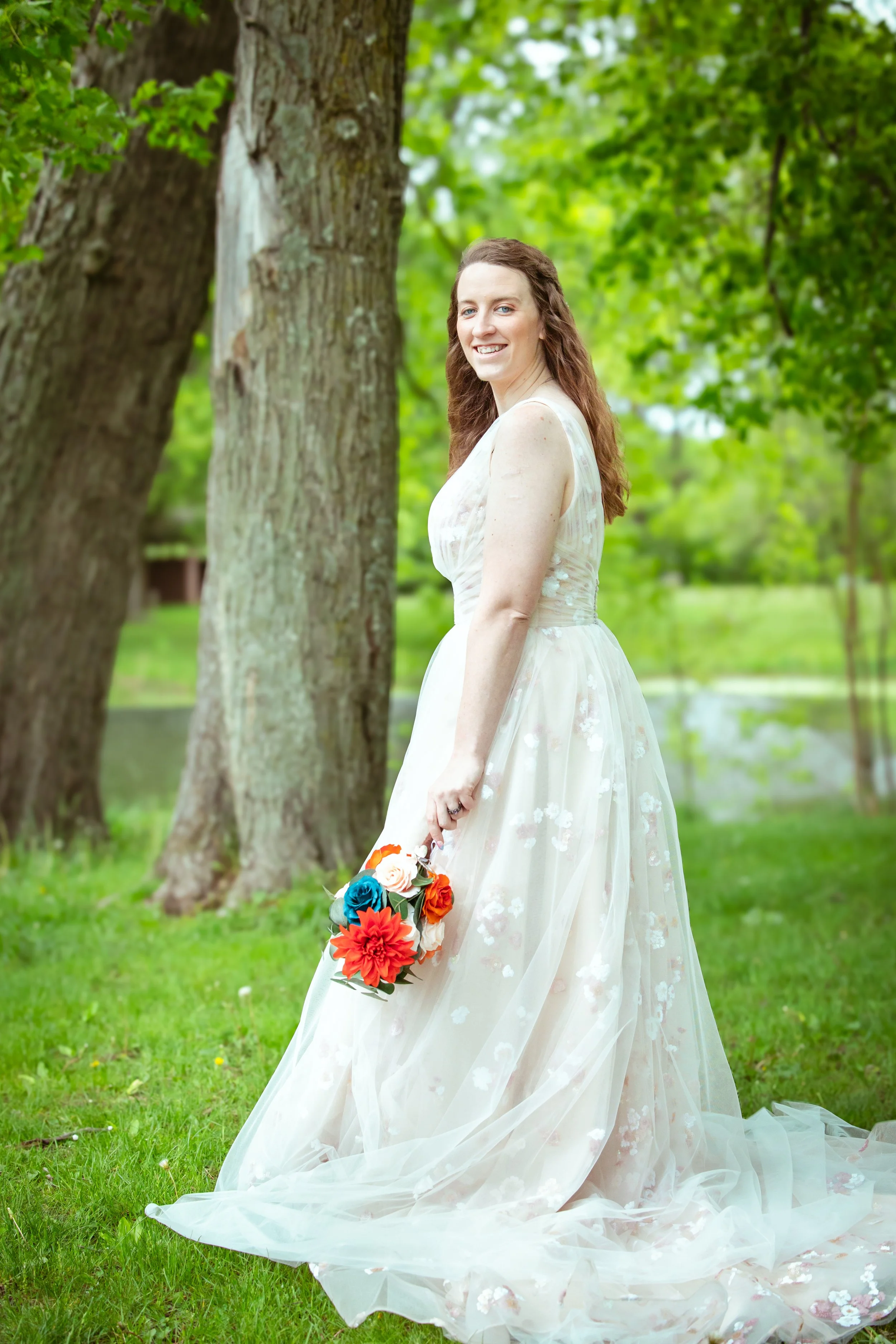 A woman in a light-colored floral wedding dress holding a bouquet of orange, red, blue, and white flowers, standing outdoors on green grass near trees.