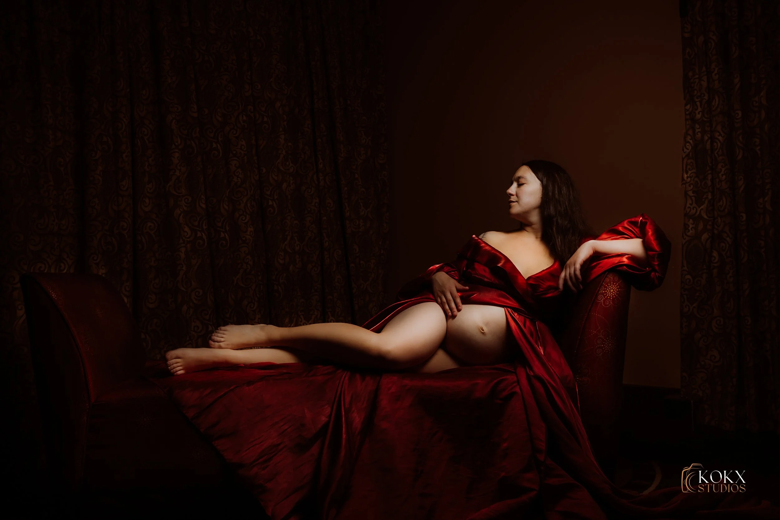 A woman in a red satin dress reclining on a bed with dark red sheets, against a dark brown background, with closed eyes and relaxed pose.