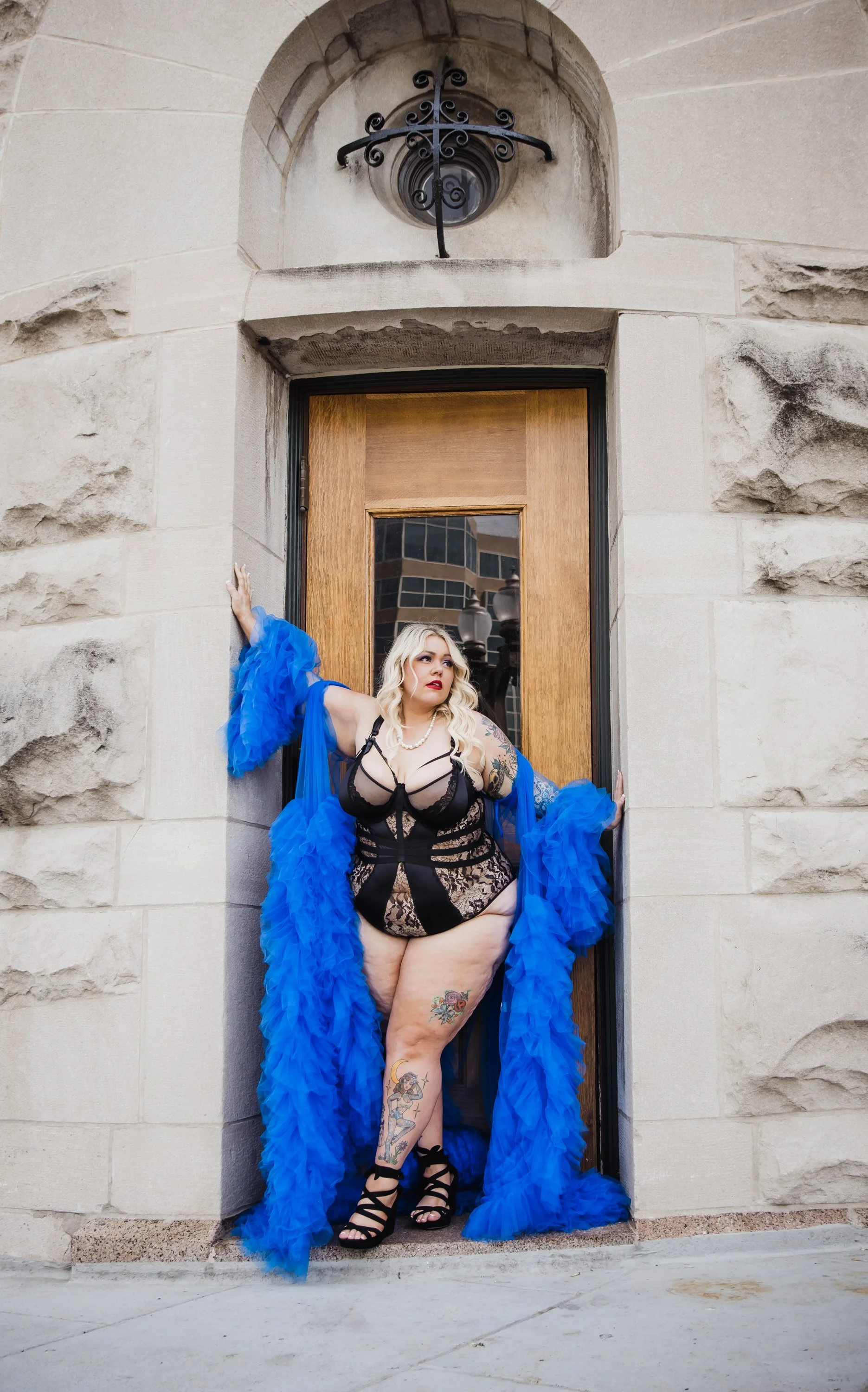 A woman with blonde hair, tattoos, and red lipstick stands in a doorway wearing black lace lingerie and a blue feathered robe, with black high heels, against a stone building exterior.