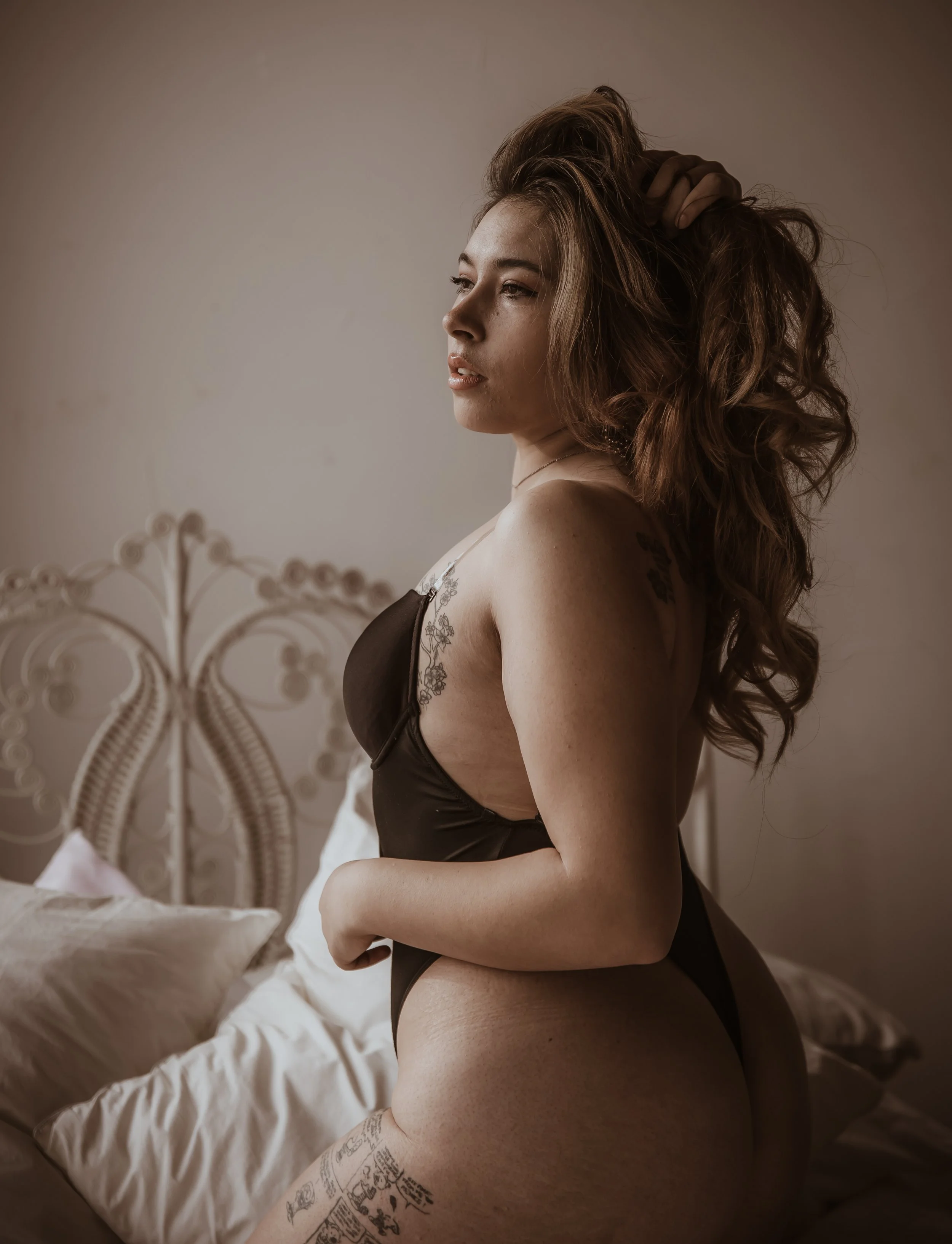 A woman with long curly hair and tattoos on her shoulder and thigh, wearing black lingerie, sitting on a bed and looking thoughtfully to the side in a softly lit room.