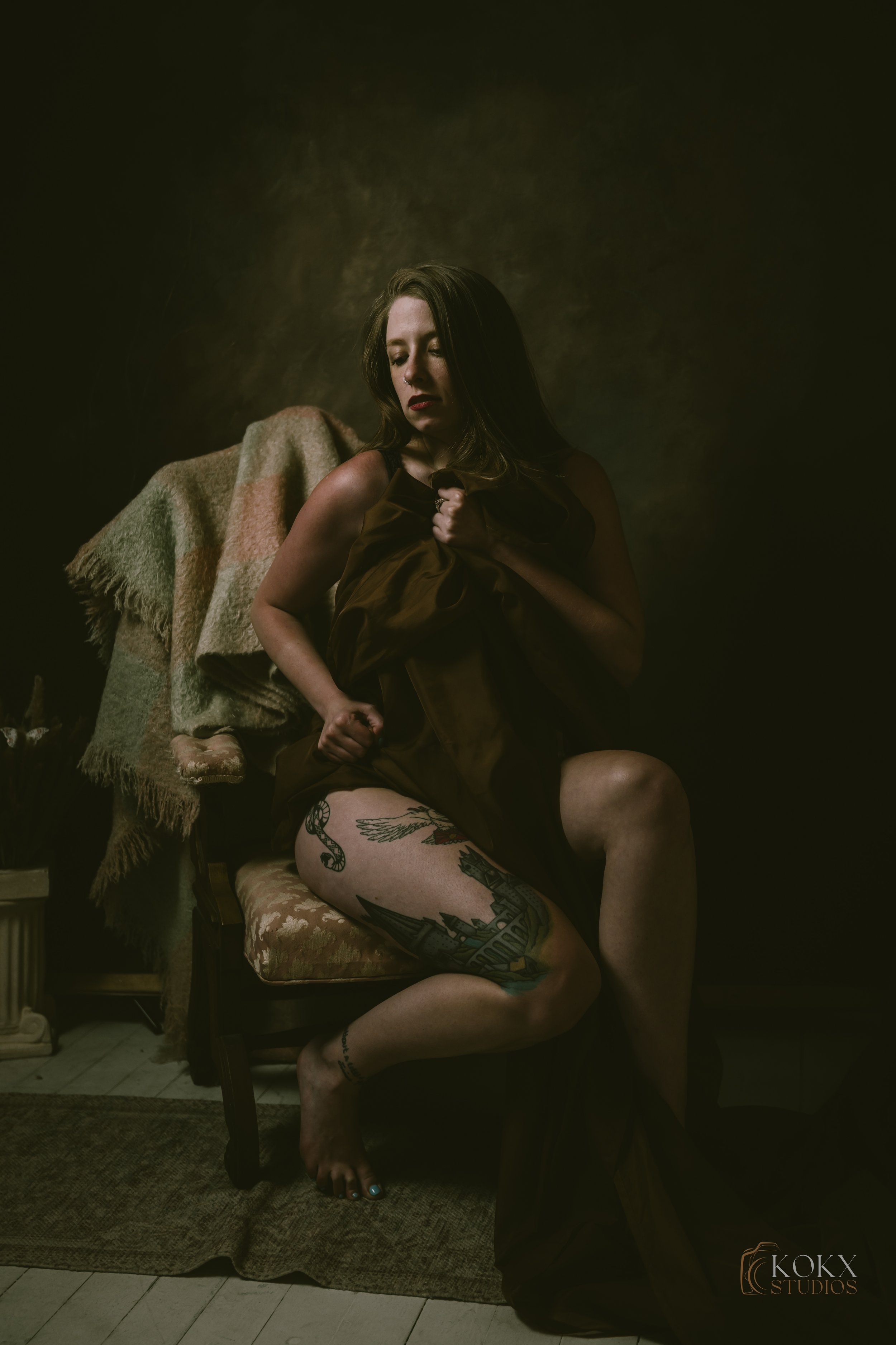 A woman with tattoos seated on an antique chair in a dimly lit room, holding a fabric close to her chest. She has shoulder-length hair and is wearing dark makeup, with a blanket draped over the back of the chair and a textured wall in the background.
