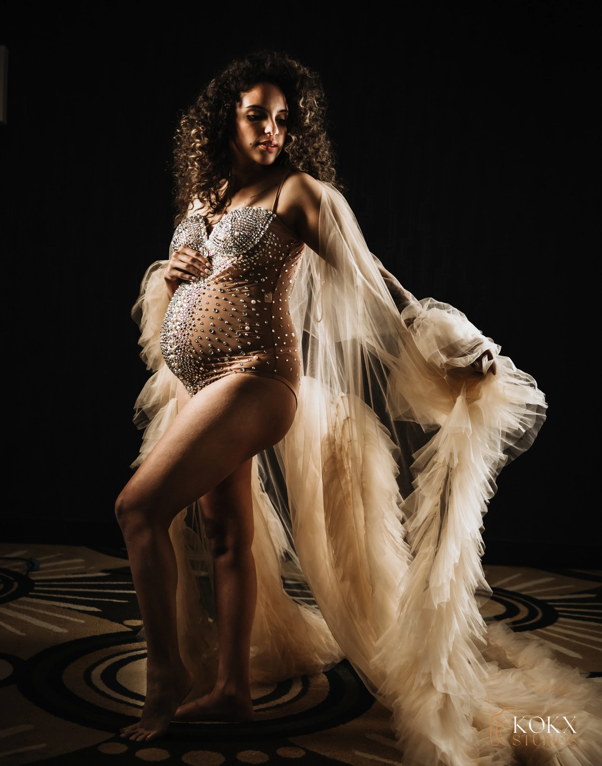 A pregnant woman in a bejeweled, sheer nude-colored dress with tulle sleeves poses indoors against a dark background.