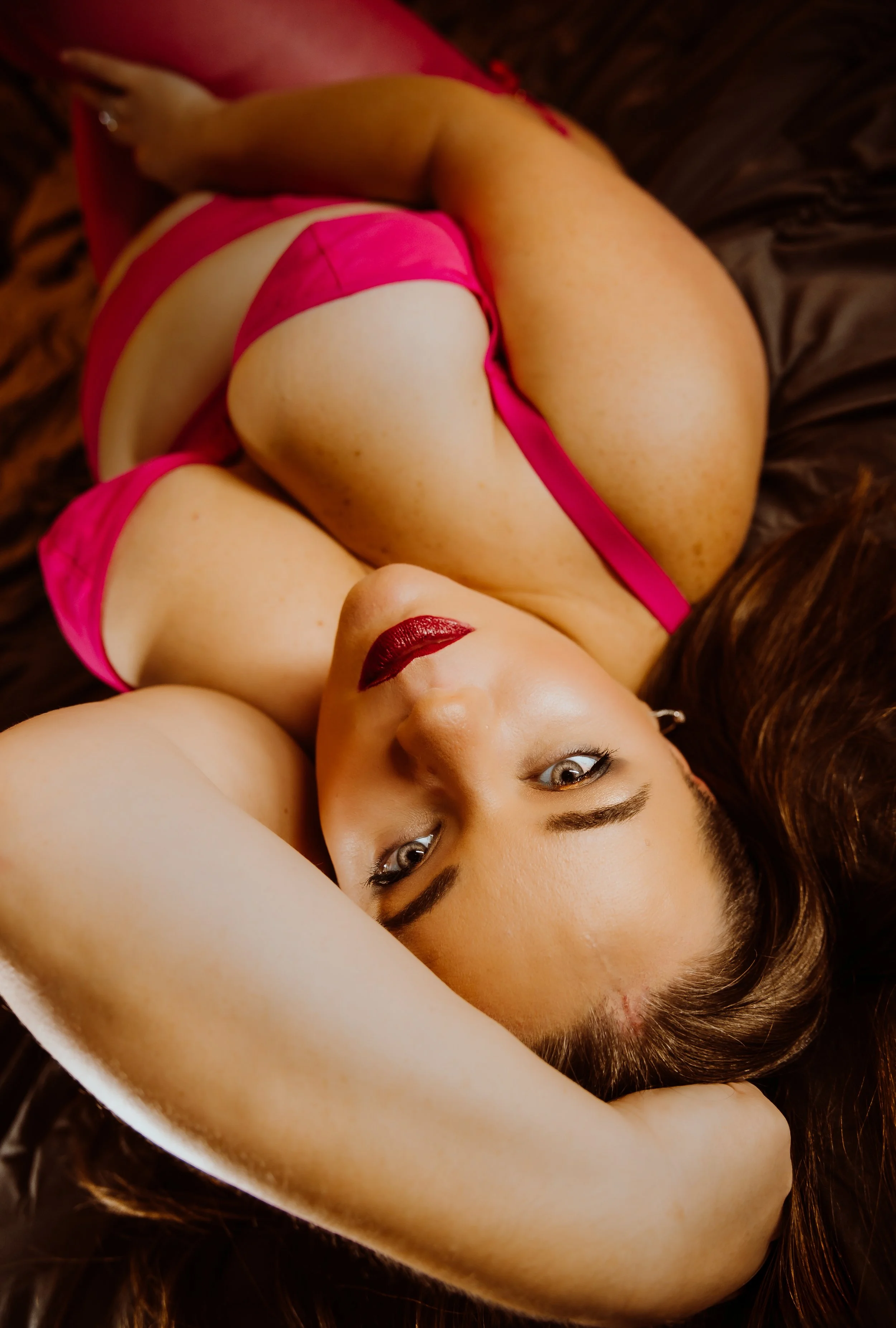 A woman with long brown hair and red lipstick is lying on a dark surface, wearing pink lingerie with her arm behind her head.
