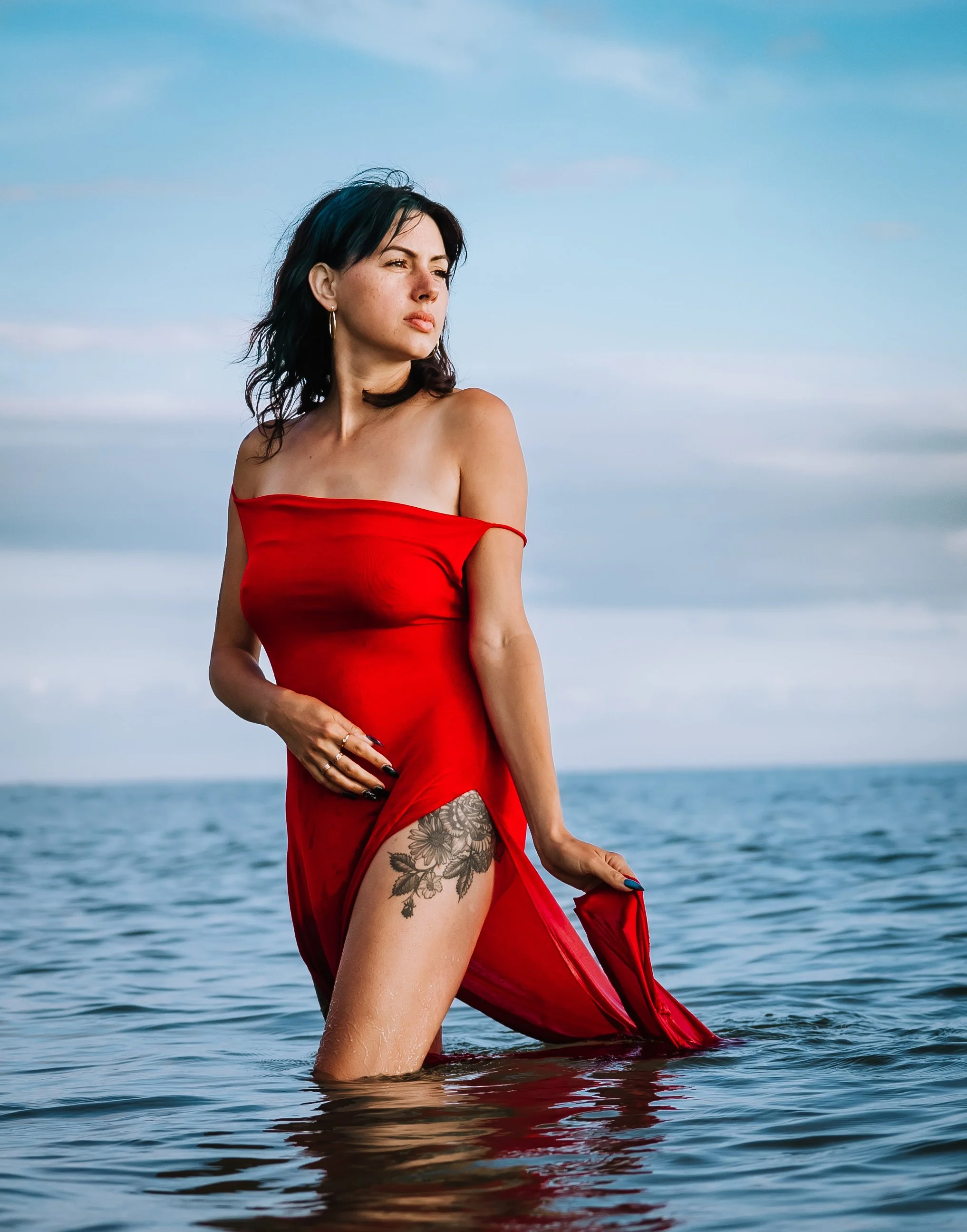 Woman in a red dress standing in the ocean with a cloudy sky background.