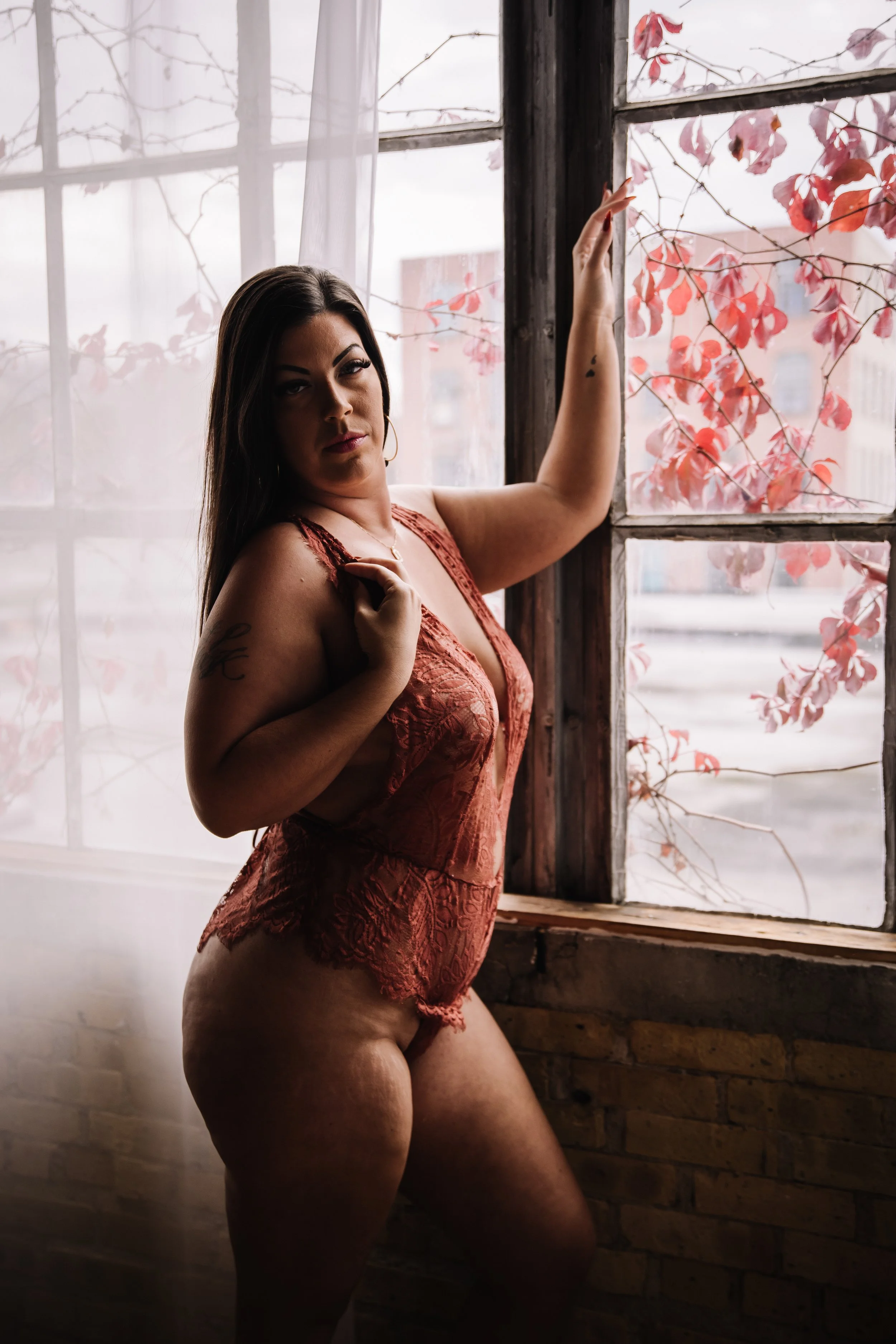 A woman standing by a rustic window with pink leaves outside, wearing a reddish lace lingerie, posing with her hand on the window frame and looking at the camera.