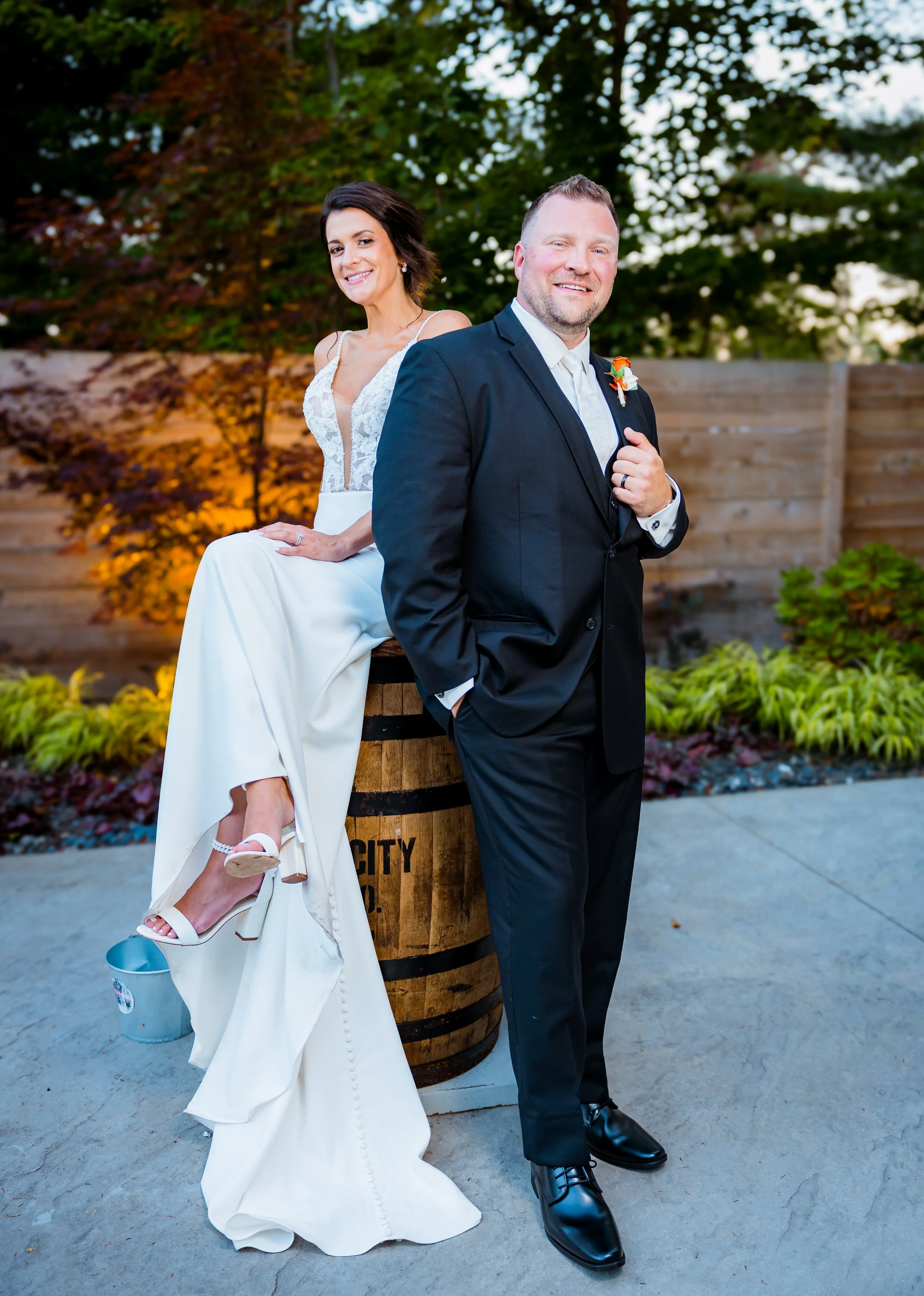 A bride in a white wedding gown and heels, sitting on a barrel, and a groom in a black suit, standing outside near a garden with trees and a wooden fence, during sunset or evening.