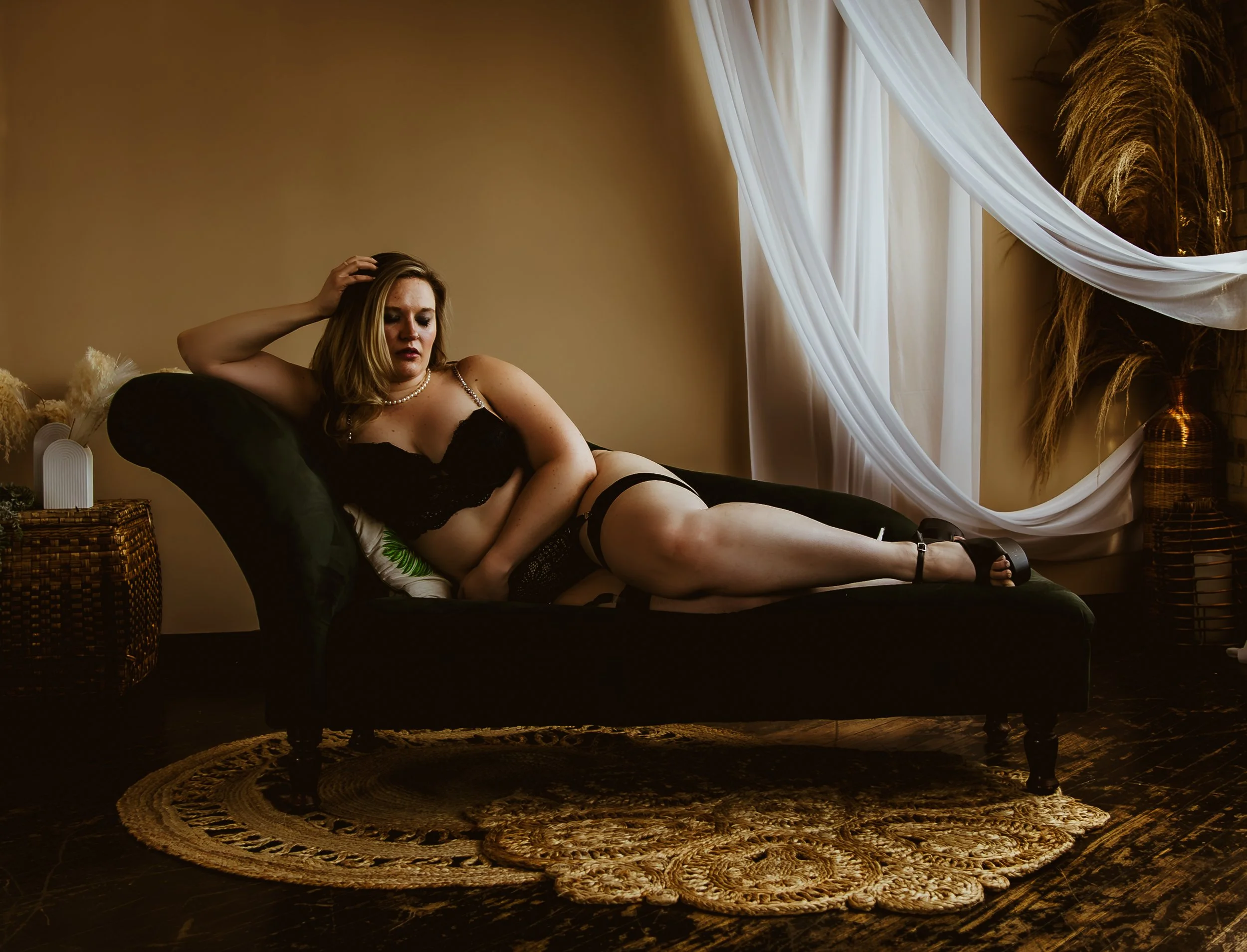 A woman in black lingerie lying on a dark green velvet chaise lounge in a warmly lit room, with beige walls, white draped curtains, and decorative vases, plants, and dried pampas grass in the background.