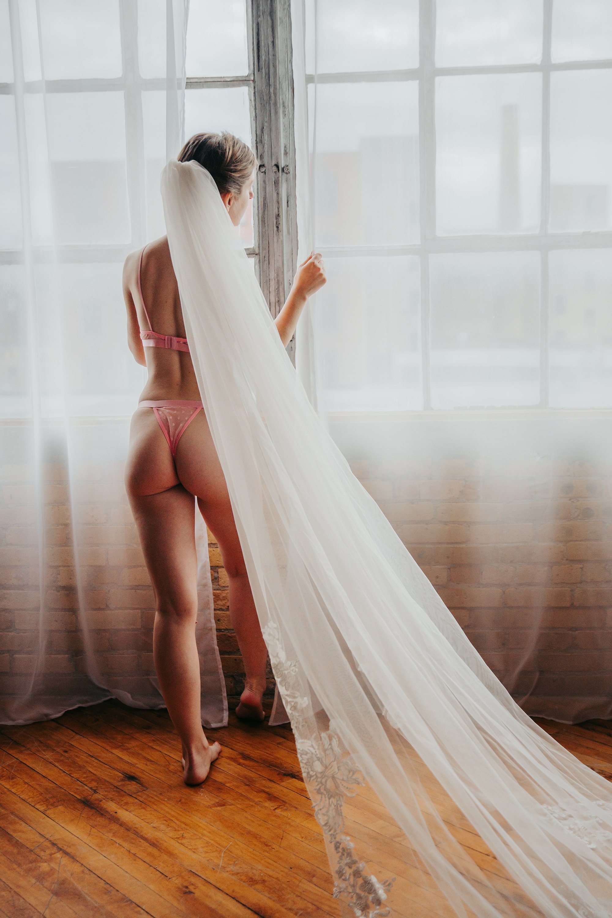 A woman in pink lingerie standing near large windows with sheer curtains, gazing outside.