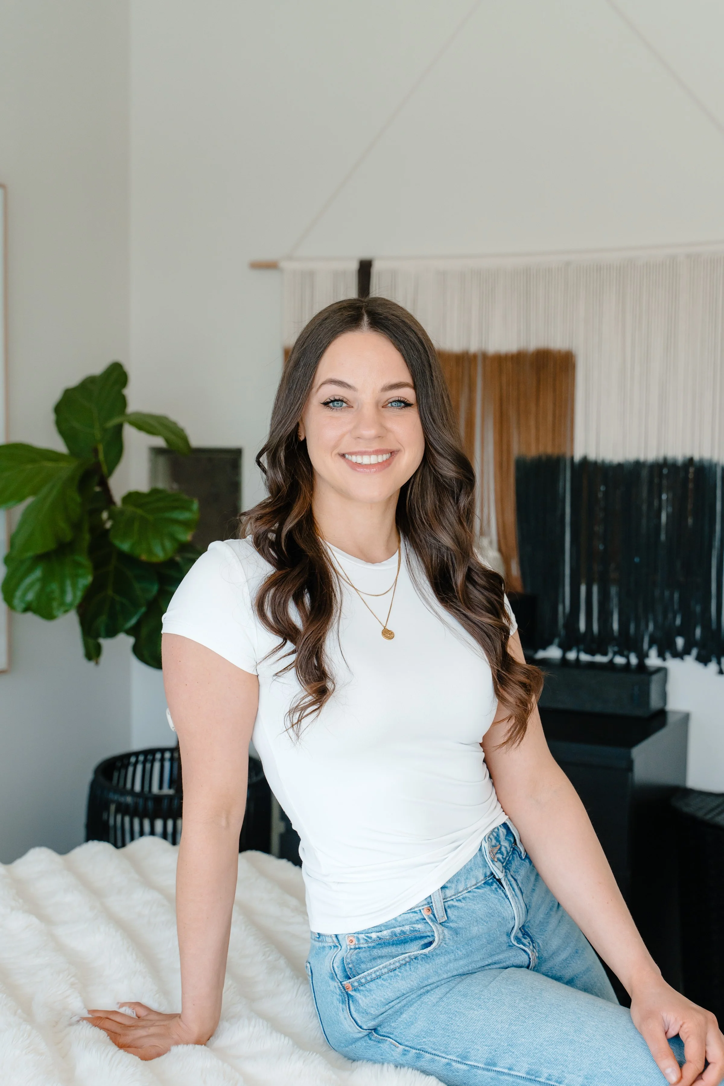 A young woman with long wavy brown hair, blue eyes, and light skin sitting on a white furry bedspread, smiling, wearing a white t-shirt, light blue jeans, and layered gold necklaces, in a modern, well-decorated room with a large green plant and a black-and-wood wall hanging in the background.