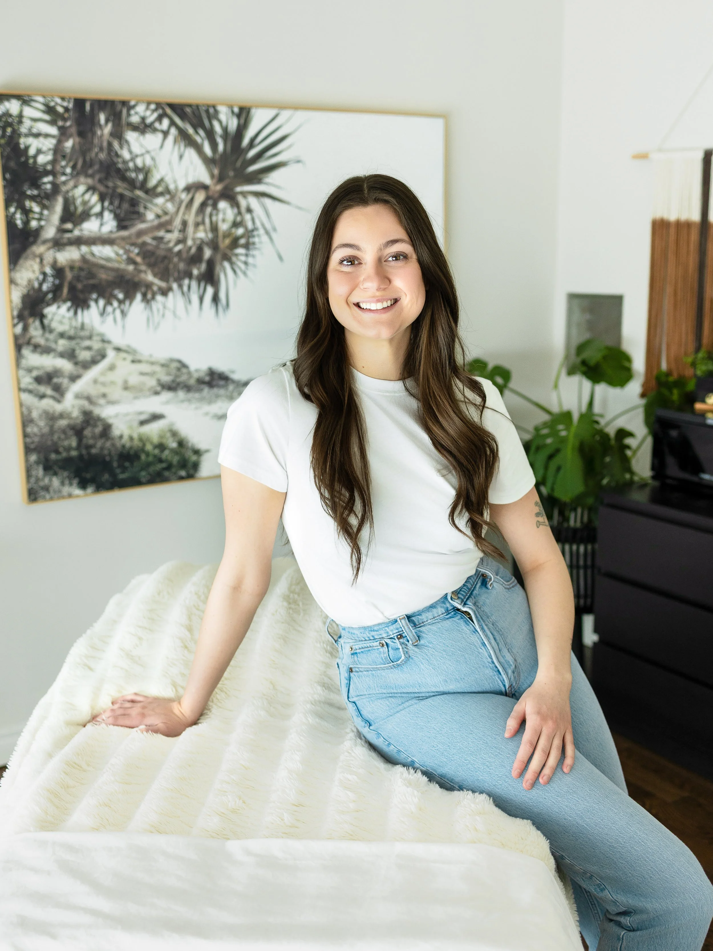 A young woman with long brown hair, wearing a white t-shirt and light blue jeans, sitting on a bed with a plush white blanket. She is smiling at the camera in a bright, modern bedroom with artwork and houseplants in the background.
