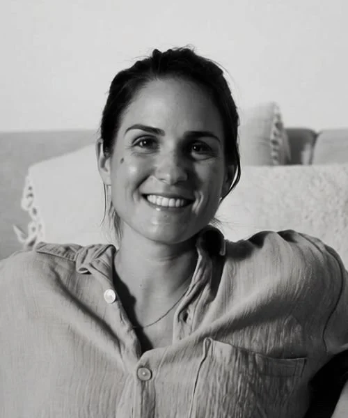 A woman with dark hair smiling, wearing a button-up shirt, sitting on a couch in a cozy living room.