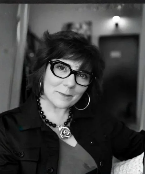 A woman with dark, wavy hair wearing glasses, hoop earrings, a necklace with a rose pendant, and a dark jacket in a black-and-white photo.