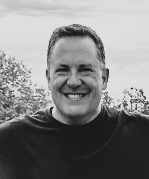 A smiling man outdoors with trees and a cloudy sky in the background.