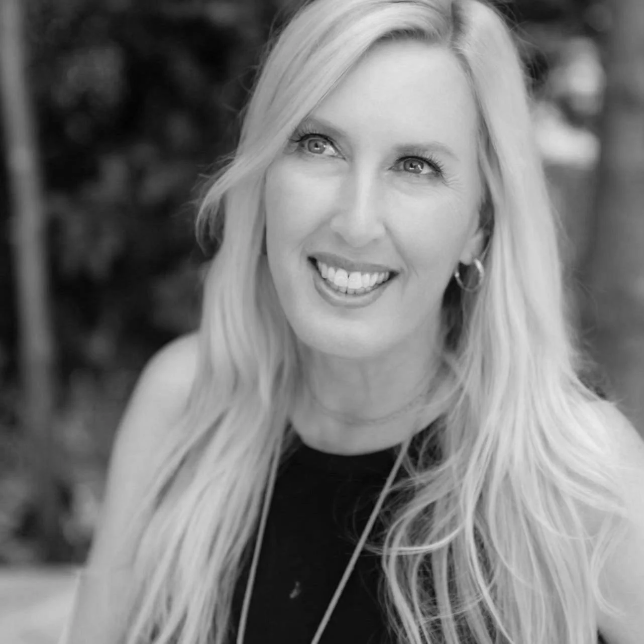 Black and white portrait of a smiling woman with long, wavy hair, wearing hoop earrings, and a dark top, outdoors with blurred foliage in the background.