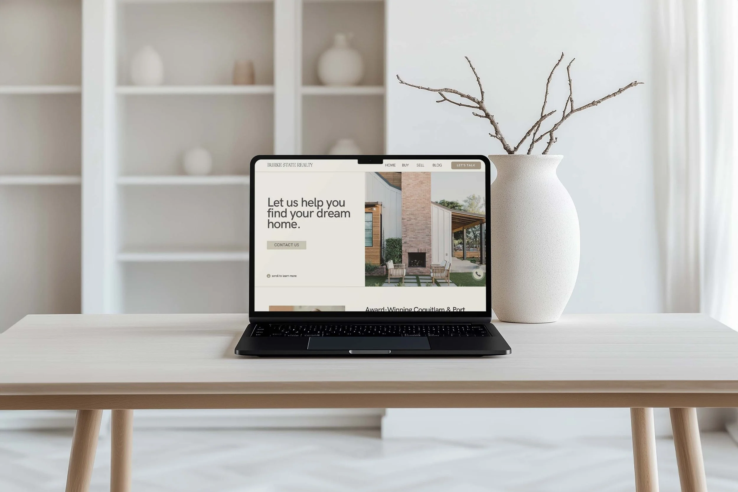 A desk with a laptop displaying a real estate website, a white textured vase with bare branches, and a white bookshelf with decorative objects in the background.