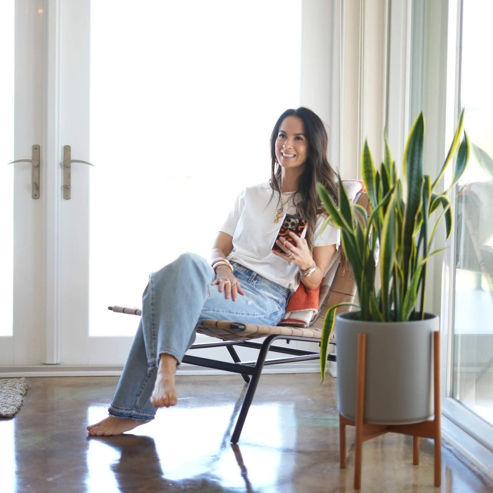Woman in a white T-shirt and light blue jeans sitting barefoot in a modern lounge chair by a bright window, smiling while holding her phone, with a tall snake plant in a gray planter beside her.