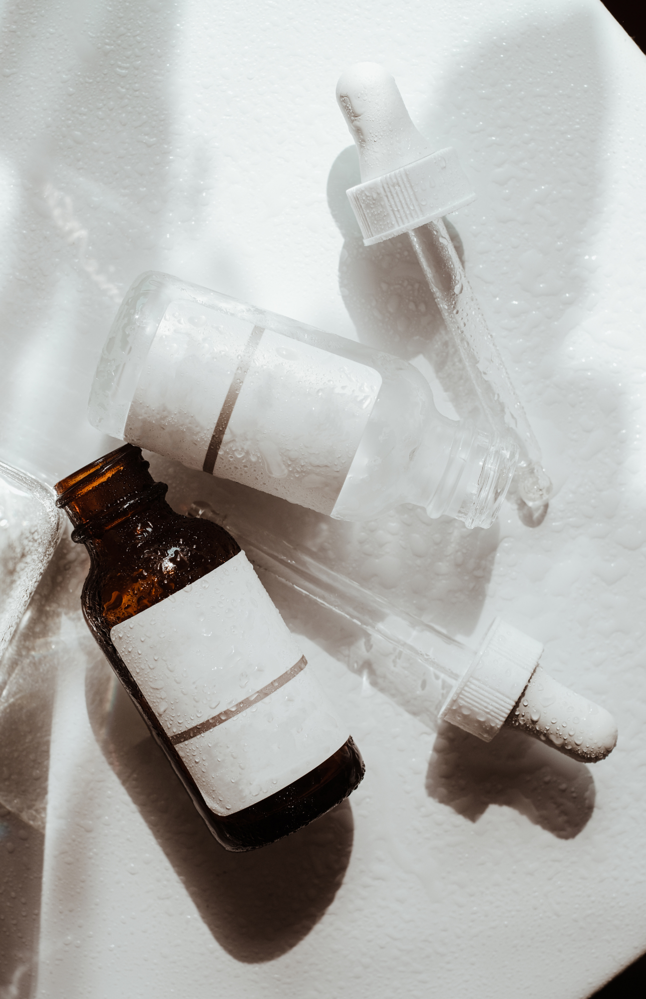 Droppers and small bottles with blank labels on a wet, illuminated surface.