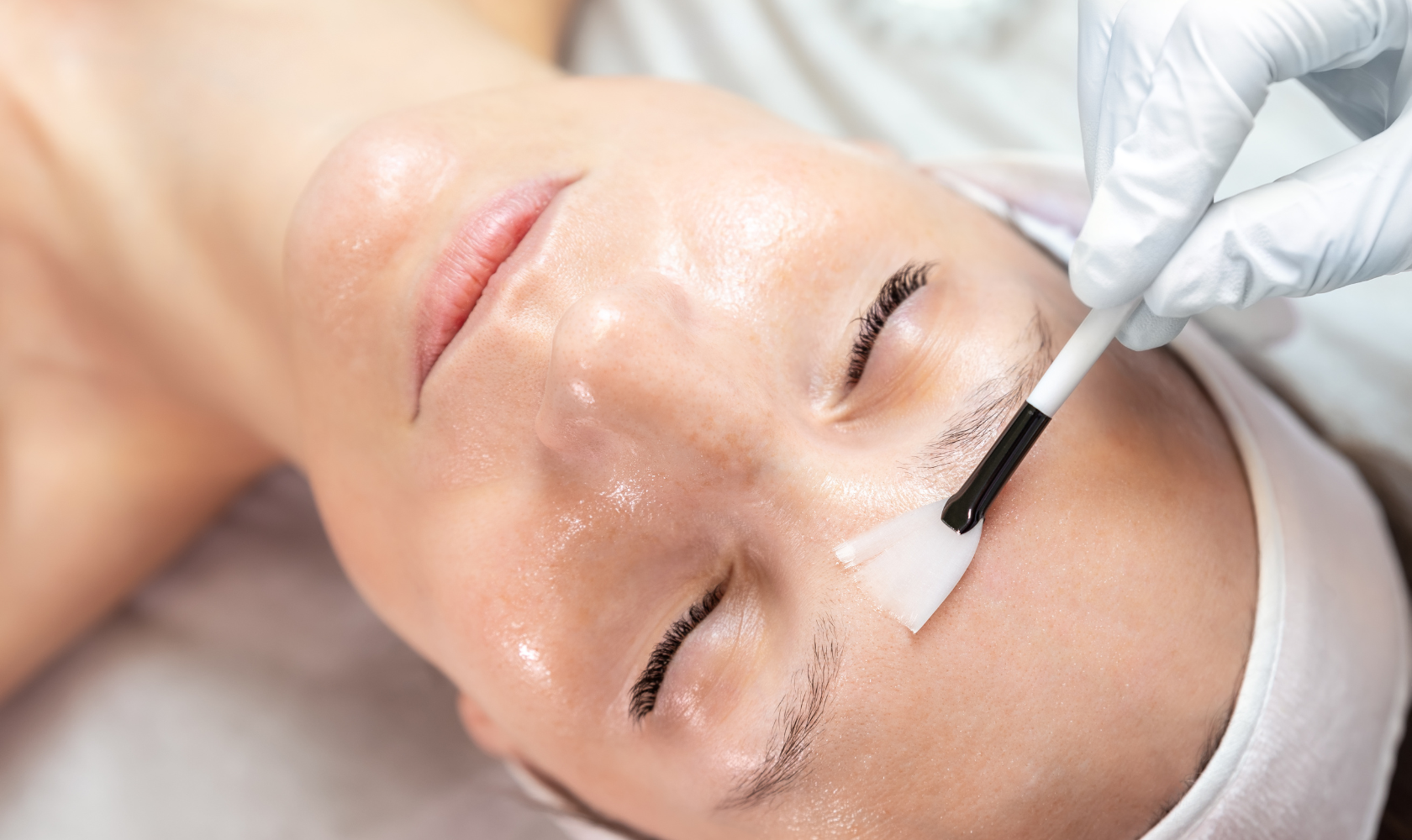 A woman receiving a facial treatment, with a skincare professional applying a product to her forehead using a brush, in a clinical setting.