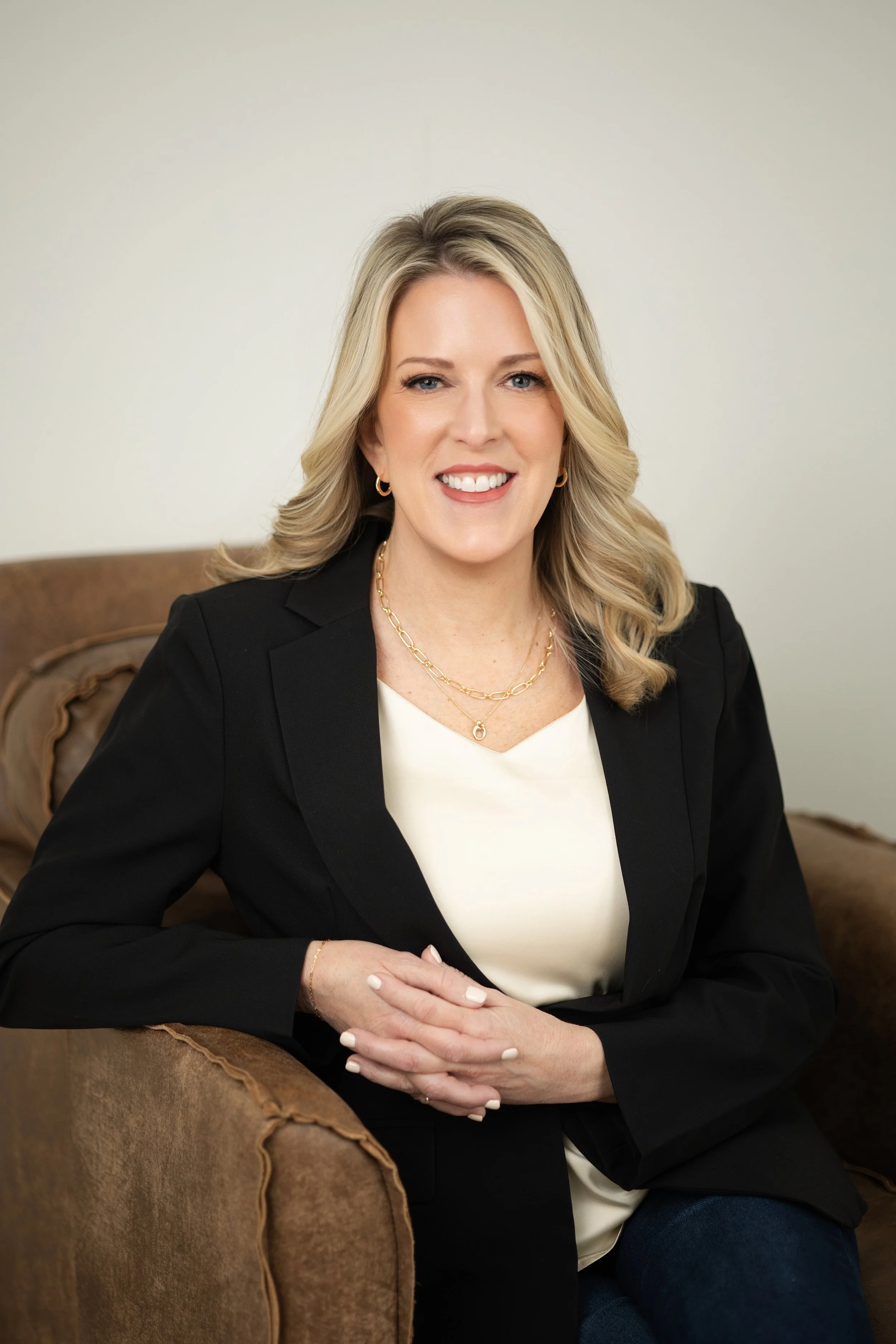 A woman with blonde hair smiling, wearing a black blazer and jewelry, sitting on a brown sofa with a cream-colored wall behind her.