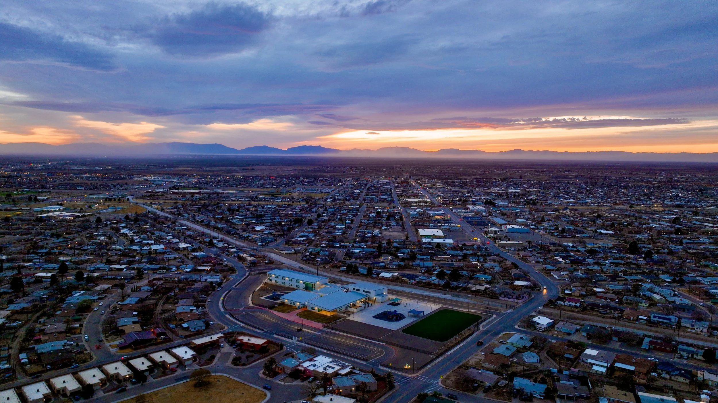 Aerial view of a cityscape at sunset, showing residential and commercial buildings with distant mountains and colorful sky.