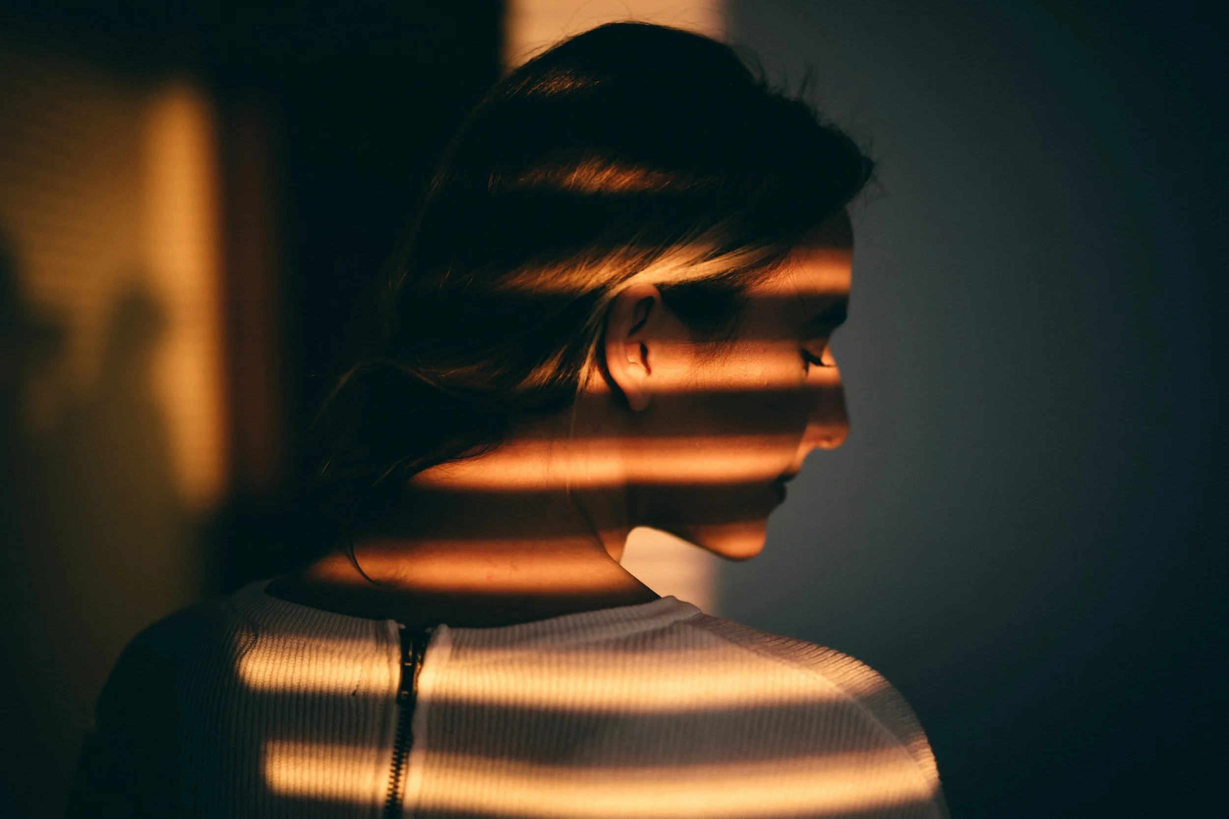A side profile of a woman with dark hair, illuminated by horizontal lines of warm light through a window in a dark room.