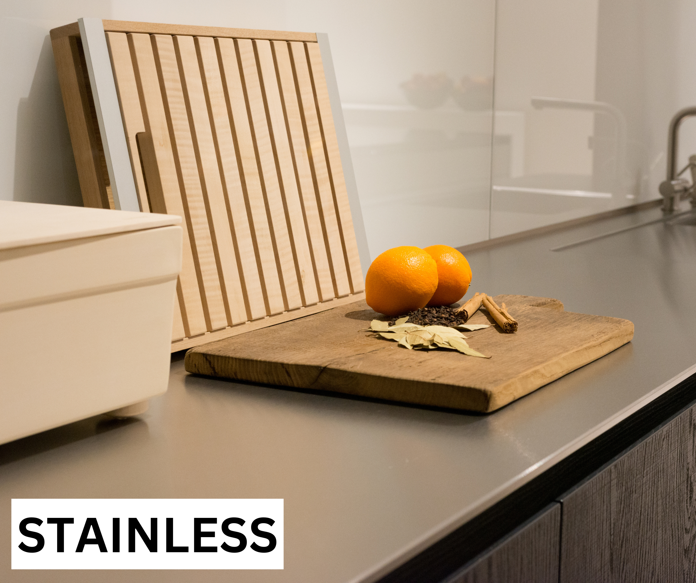 Une surface de cuisine en acier inoxydable avec un plan de découpe en bois, deux oranges, des feuilles de laurier séchées, un bâton de cannelle, et un lot d'ustensiles de cuisine en arrière-plan.