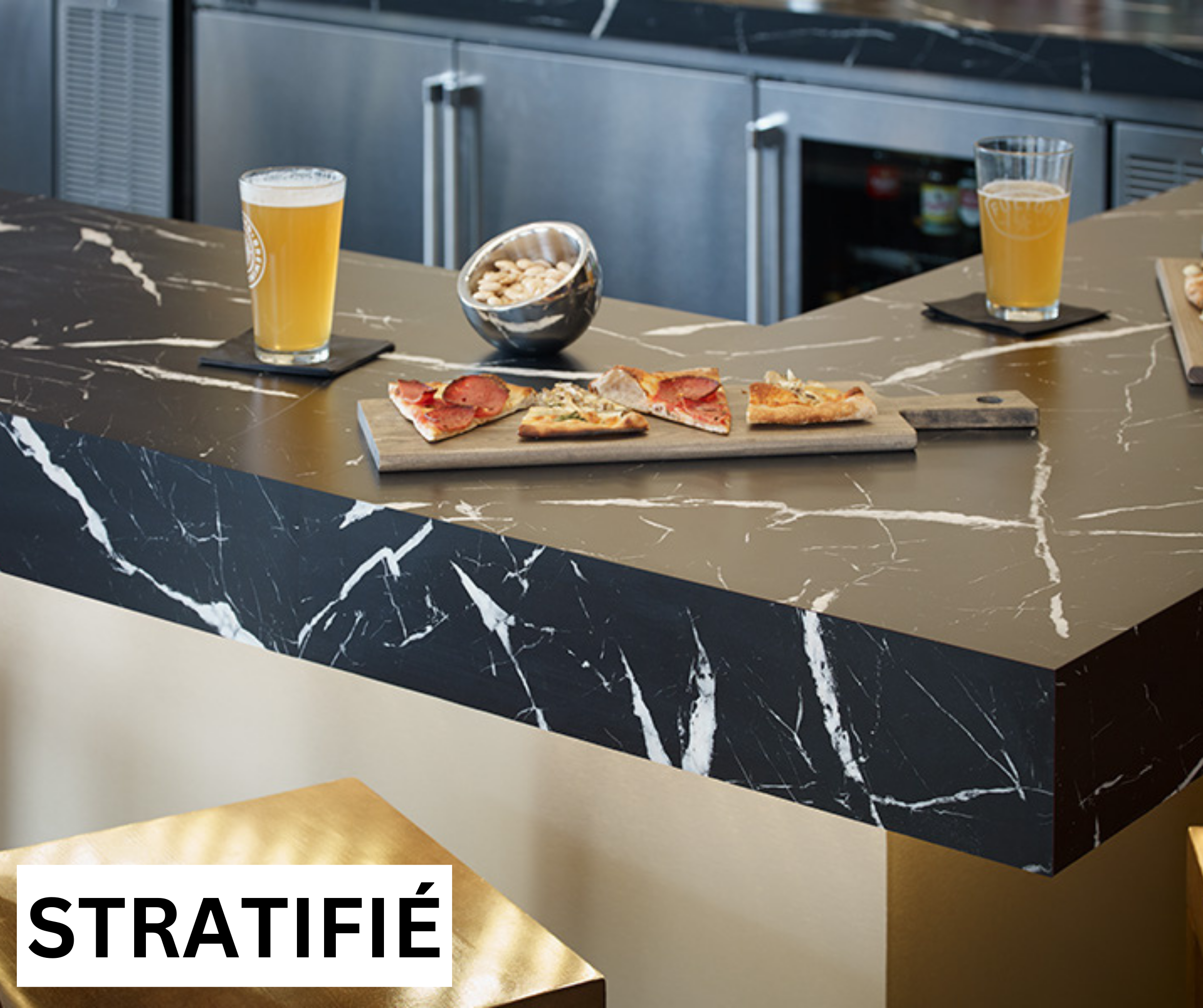 Un îlot de cuisine en pierre de lave noire avec veines blanches, avec deux verres de bière, une planche à pizza avec des parts de pizza, et un bol de noix, dans un environnement moderne.