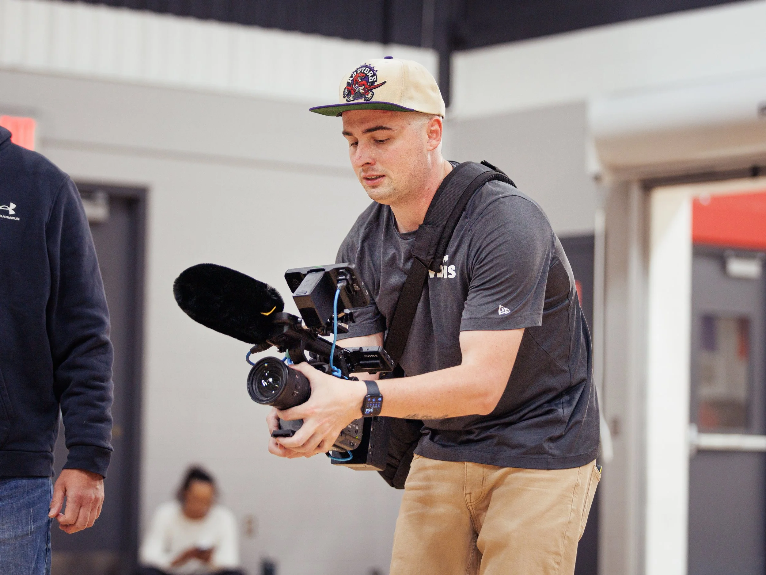 A man wearing a beige cap, black t-shirt, and khaki pants holds a professional video camera with a microphone, in an indoor setting.