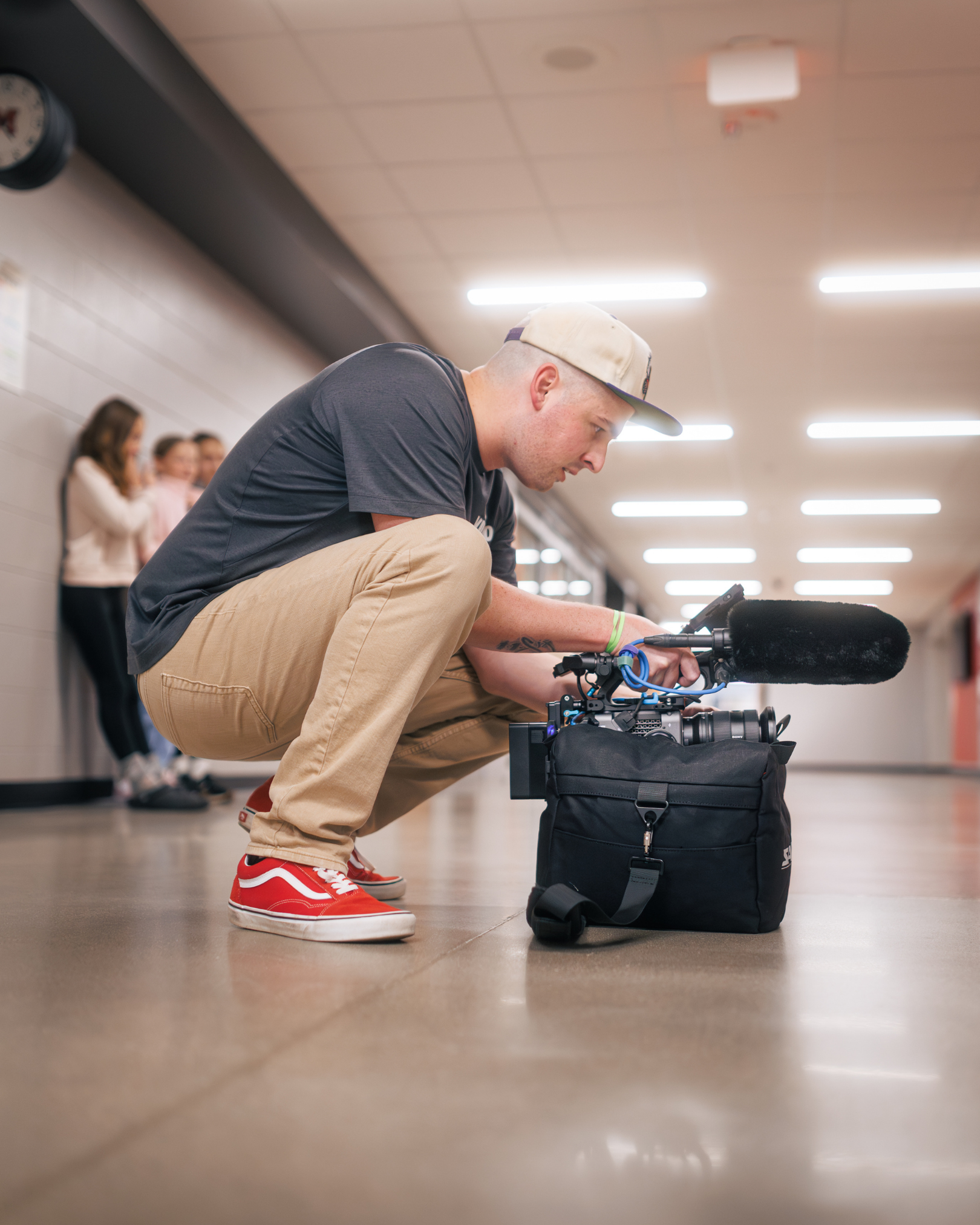 Side profile of Gavin, owner and operator of Gavin (Bison) Media, crouched low in a hallway while preparing a professional video camera for a shoot with three actresses in the background.