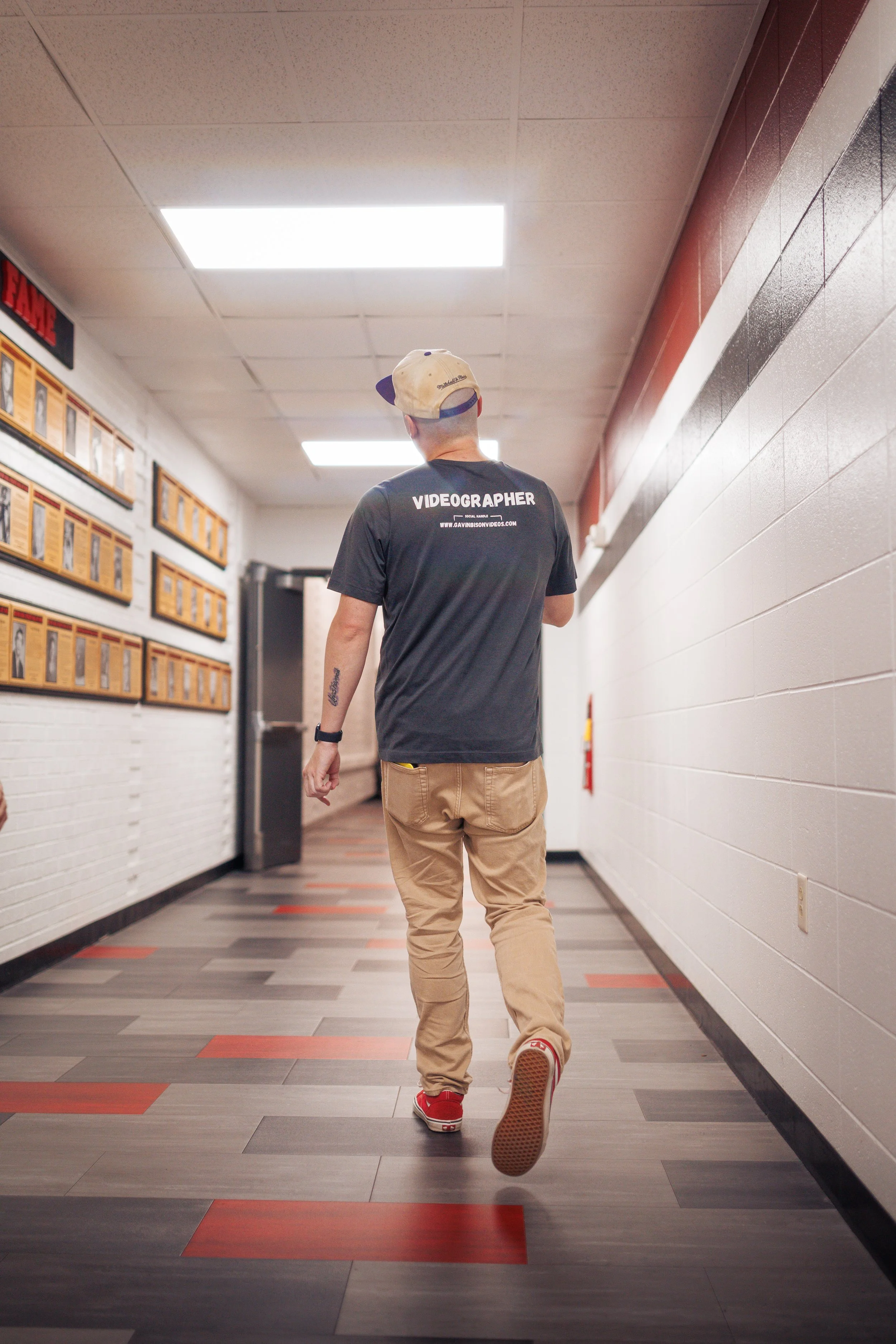 The owner and operator of Gavin (Bison) Videos walking down a corridor wearing a black t-shirt with 'Videographer' printed on the back, beige pants, red shoes, and a backwards baseball cap in an indoor setting.